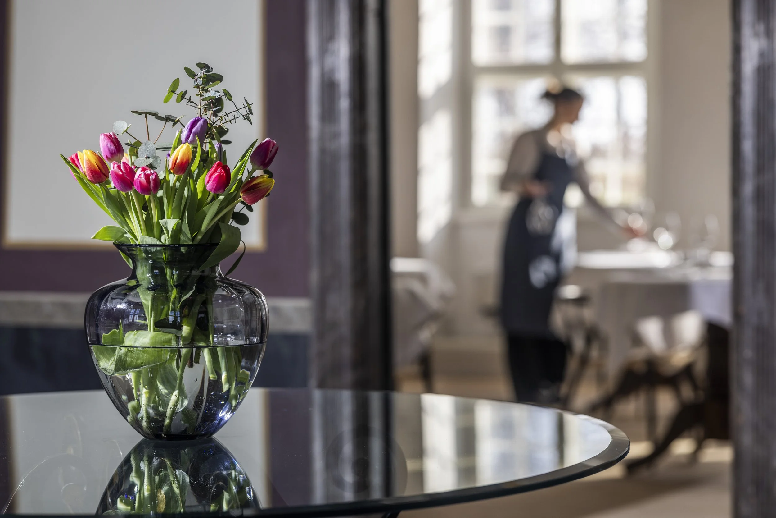 En vase med en buket tulipaner og eucalyptus grener på et rundt spejlbord, med en bagvedliggende kvinde i et lyst rum med store vinduer.