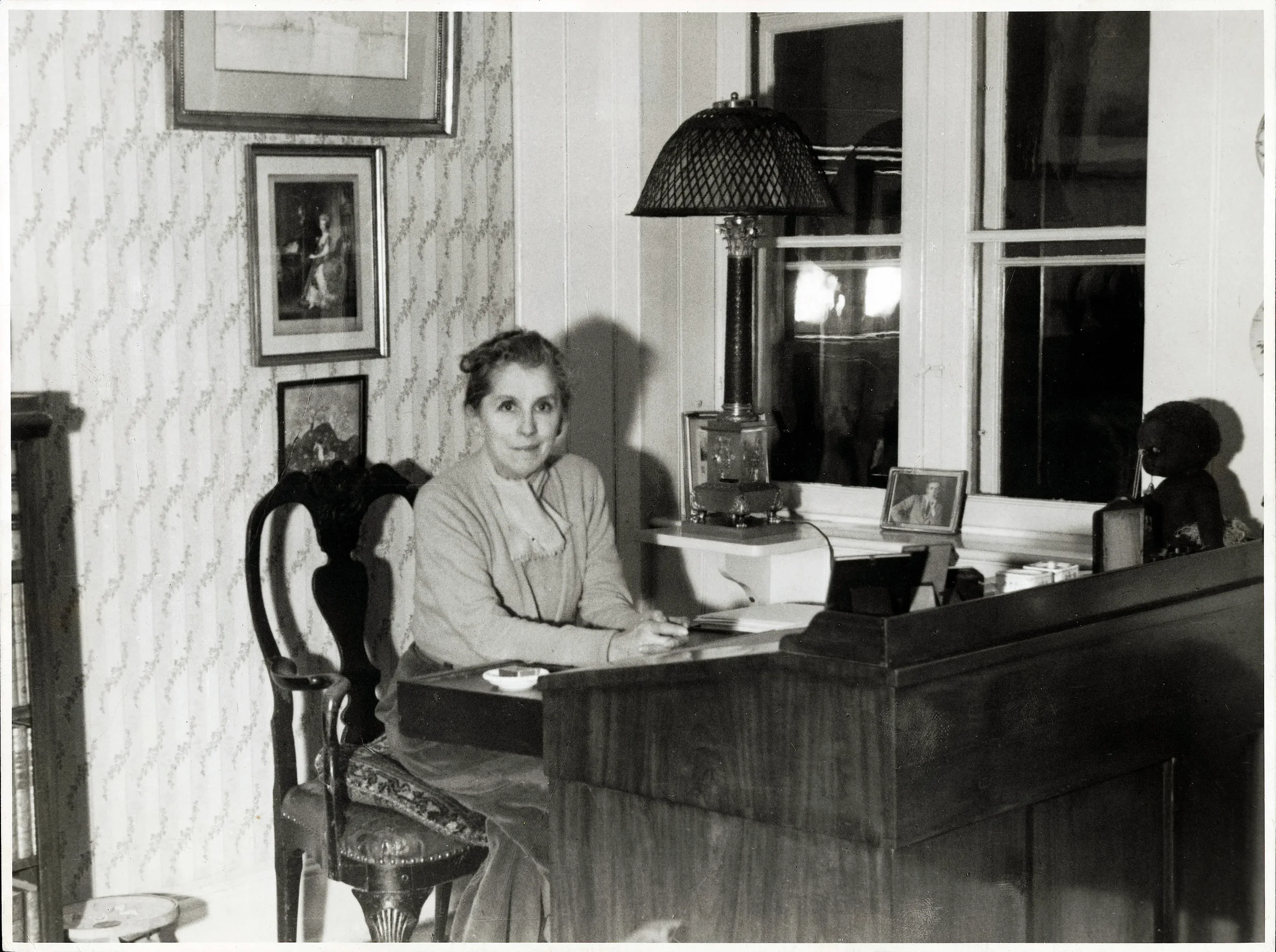 En kvinde sidder ved et klaver i et værelse med vintage-indretning, inklusive billeder på væggene og en lampe ved vinduet.