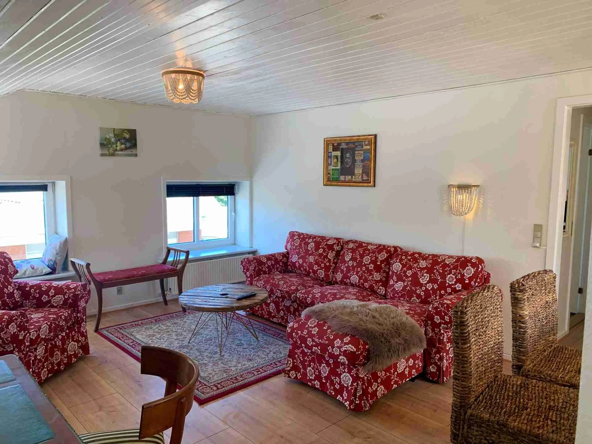 Stue med rød og hvidt mønster på sofaer og stole, træbord, tæppe, to vinduer med mørklægningsruller og vægkunst.