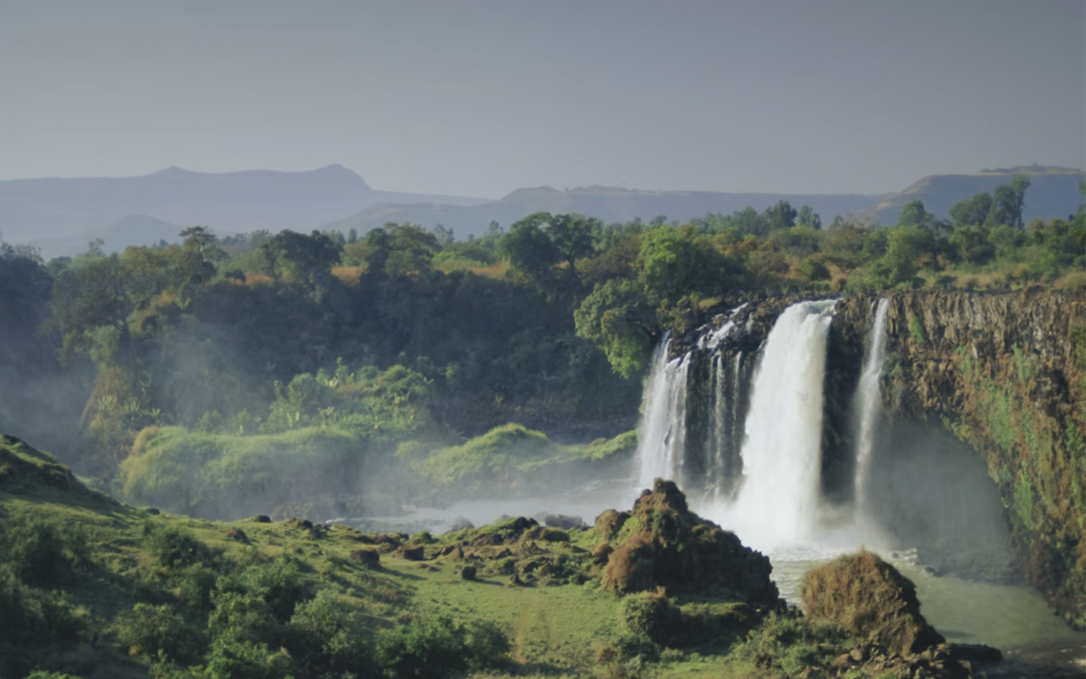 Ferskvandfald, omgivet af grønne skove og bakker under en klar himmel.