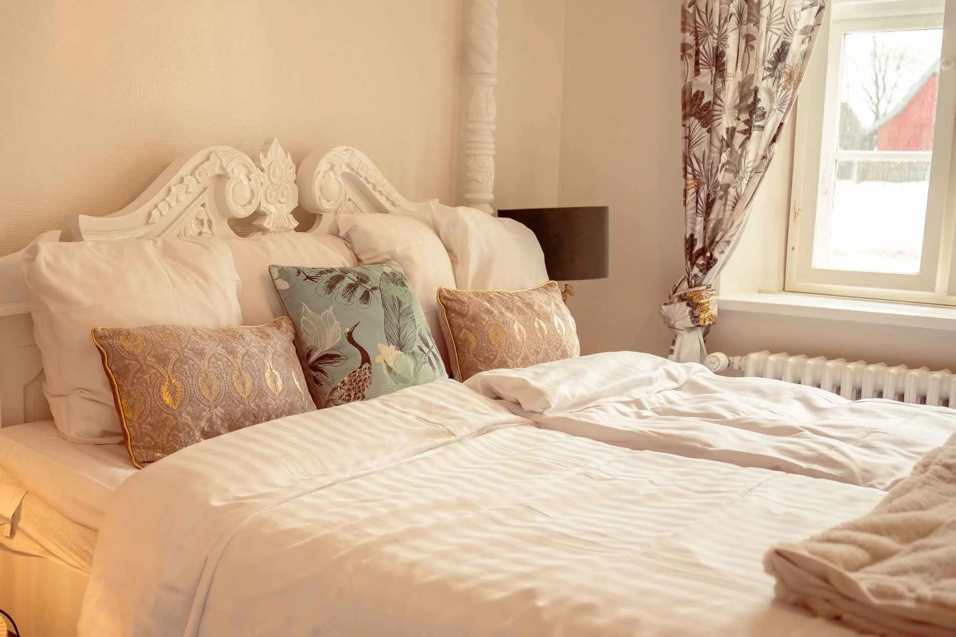 Udskåret hvid sengegavl, teal puder, pelstæppe, vindue