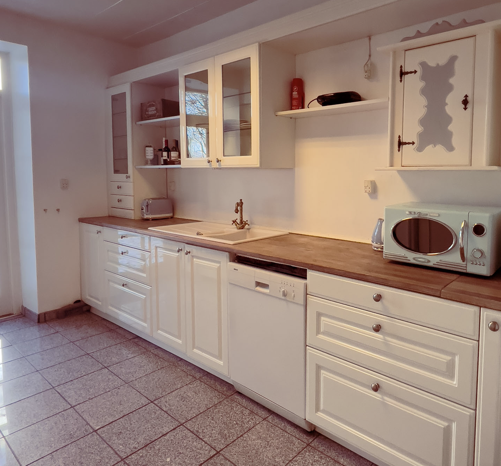 Køkken med hvide skabe, træbordplade, vask, mikroovn, opbevaringshylder, og vindue