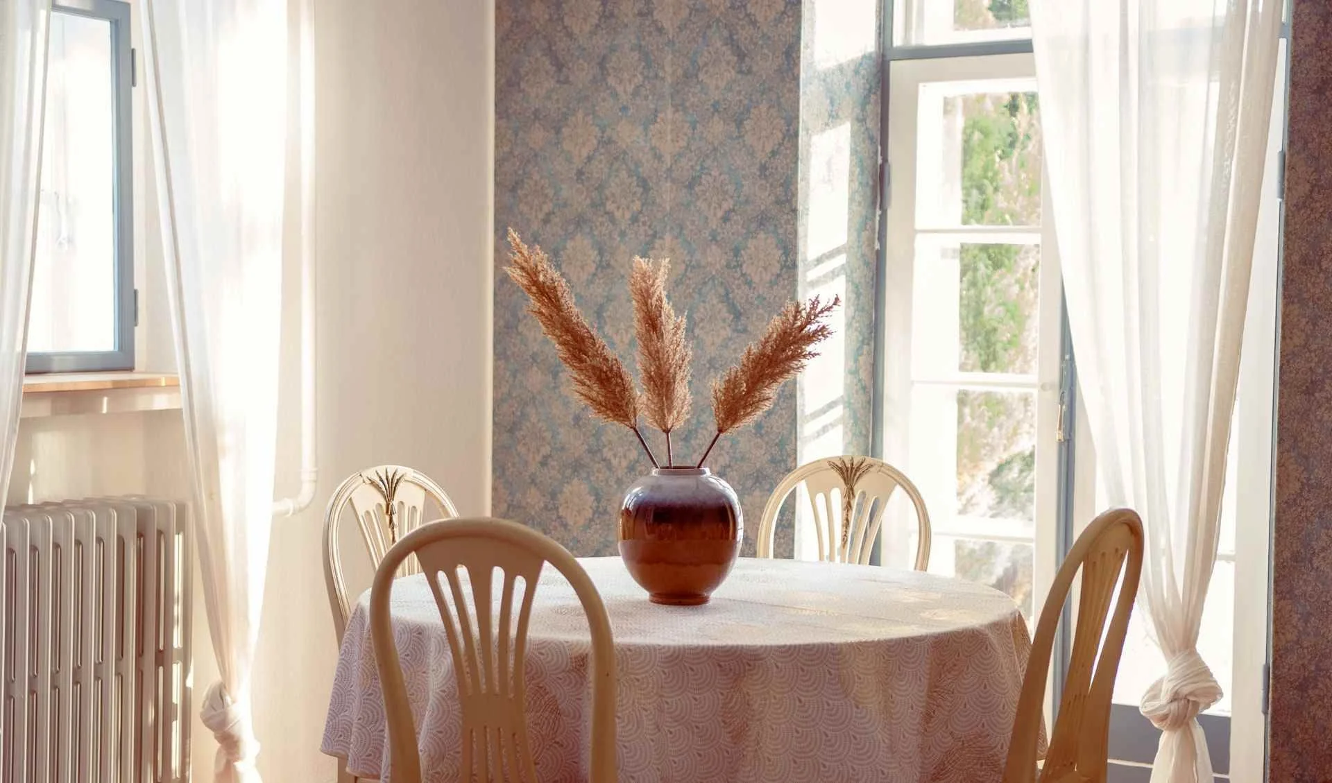Et rundt spisebord med en hvid dug og en rødbrun vase med tørrede plantegrene foran et stort vindue med hvide forhæng, i et lyst rum.