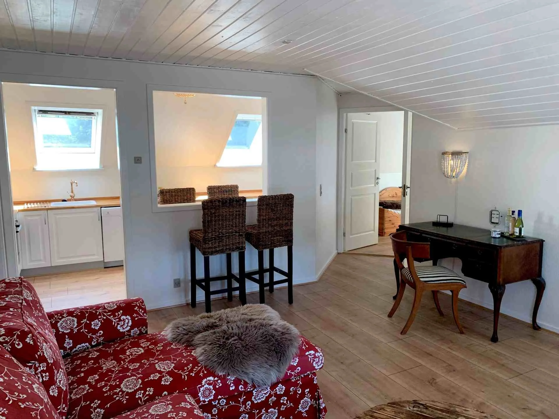 Et hyggeligt stue med en rød blomstret sofa, en lille skrivebord med et spejl og nogle alkohol flasker, en pejs og en stol, et åbent køkken med to skodder vinduer, en madras i et værelse gennem en åben dør.