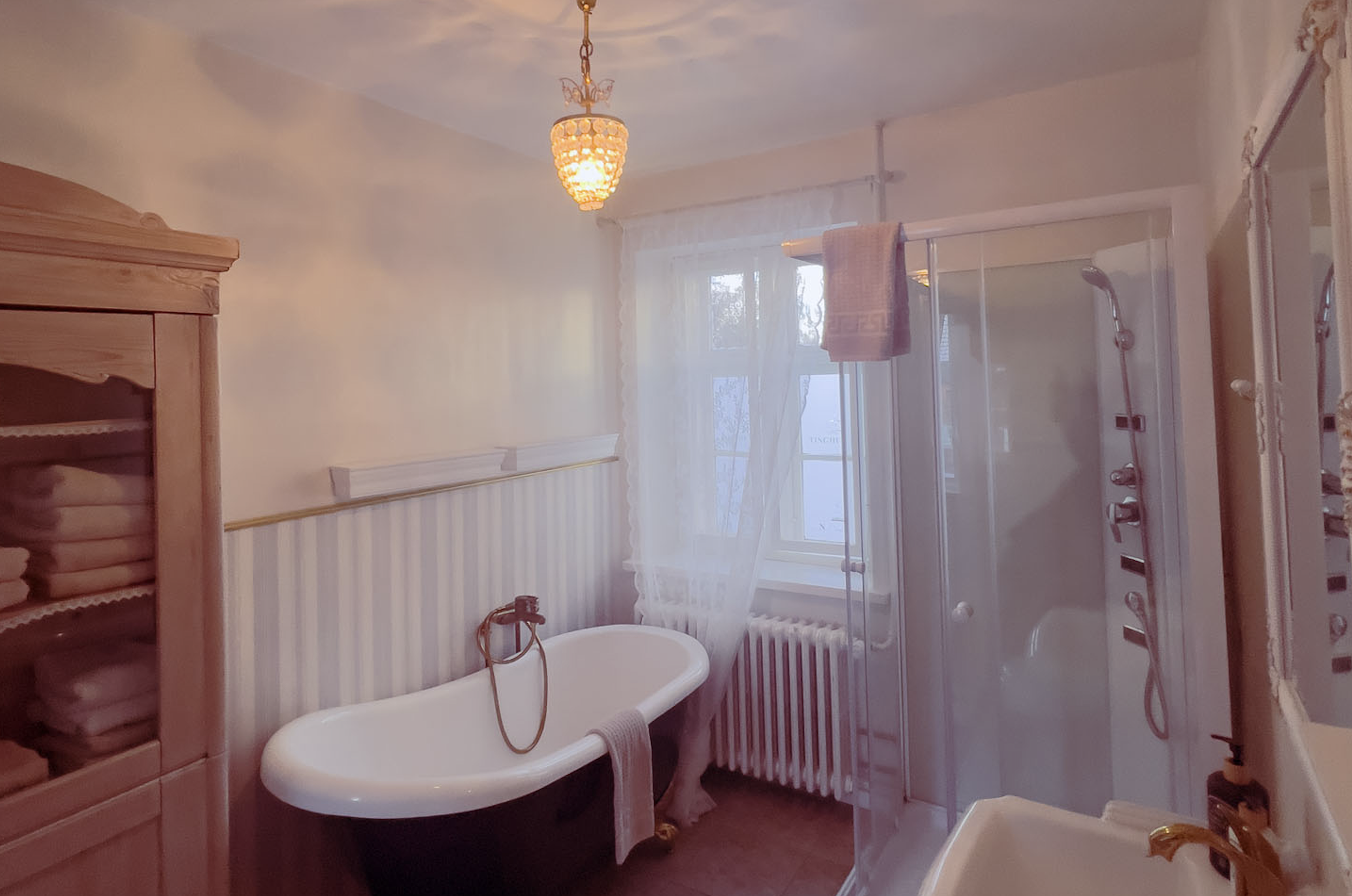 Badeværelse med badekar, glasbrusekabine, vindue med gardin, træskab med håndklæder, vask med messinghåndtag, loftlampe, stribet vægpanel.