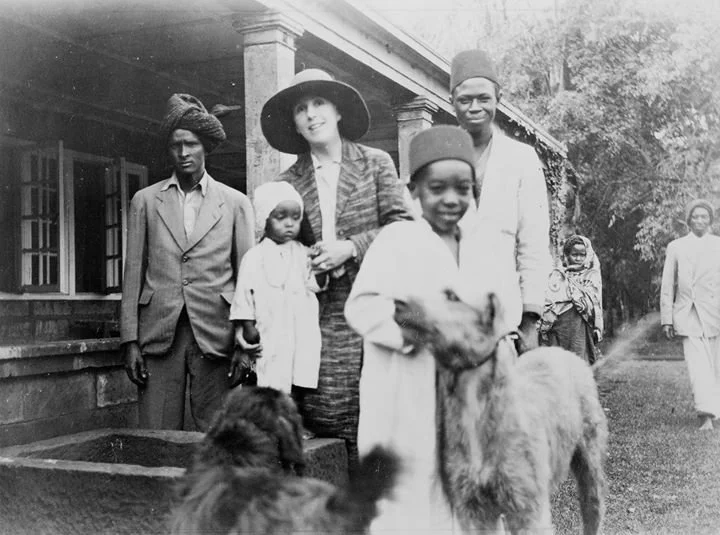 Sort/hvid foto af en gruppe mennesker udenfor en bygning. En kvinde holder en barn, hvilket også er en dreng med en ged. Flere personer står i baggrunden.