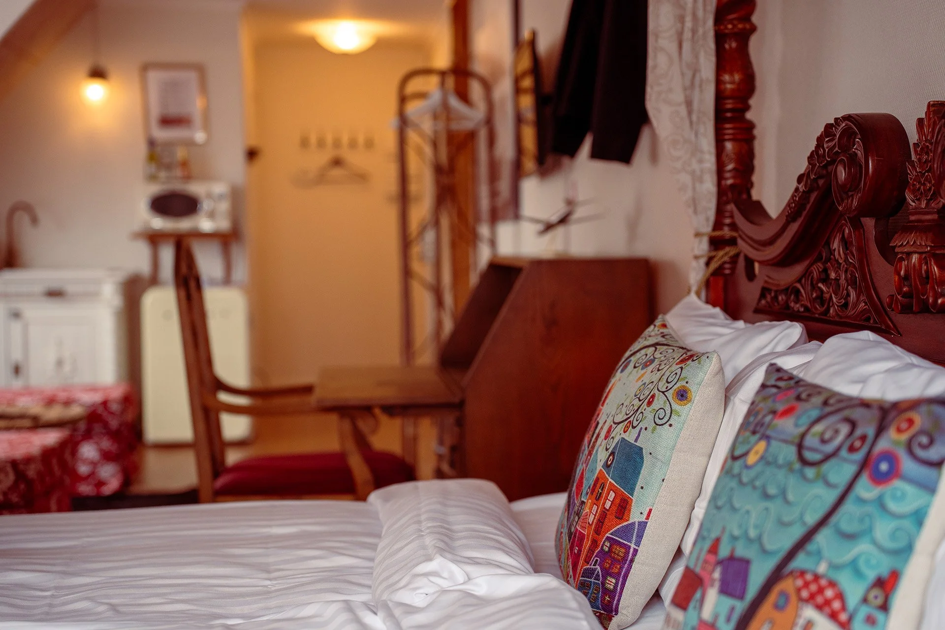 Soveværelse med en seng, dekorative puder med farverige bylandskaber, træmøbler og varme belysning.