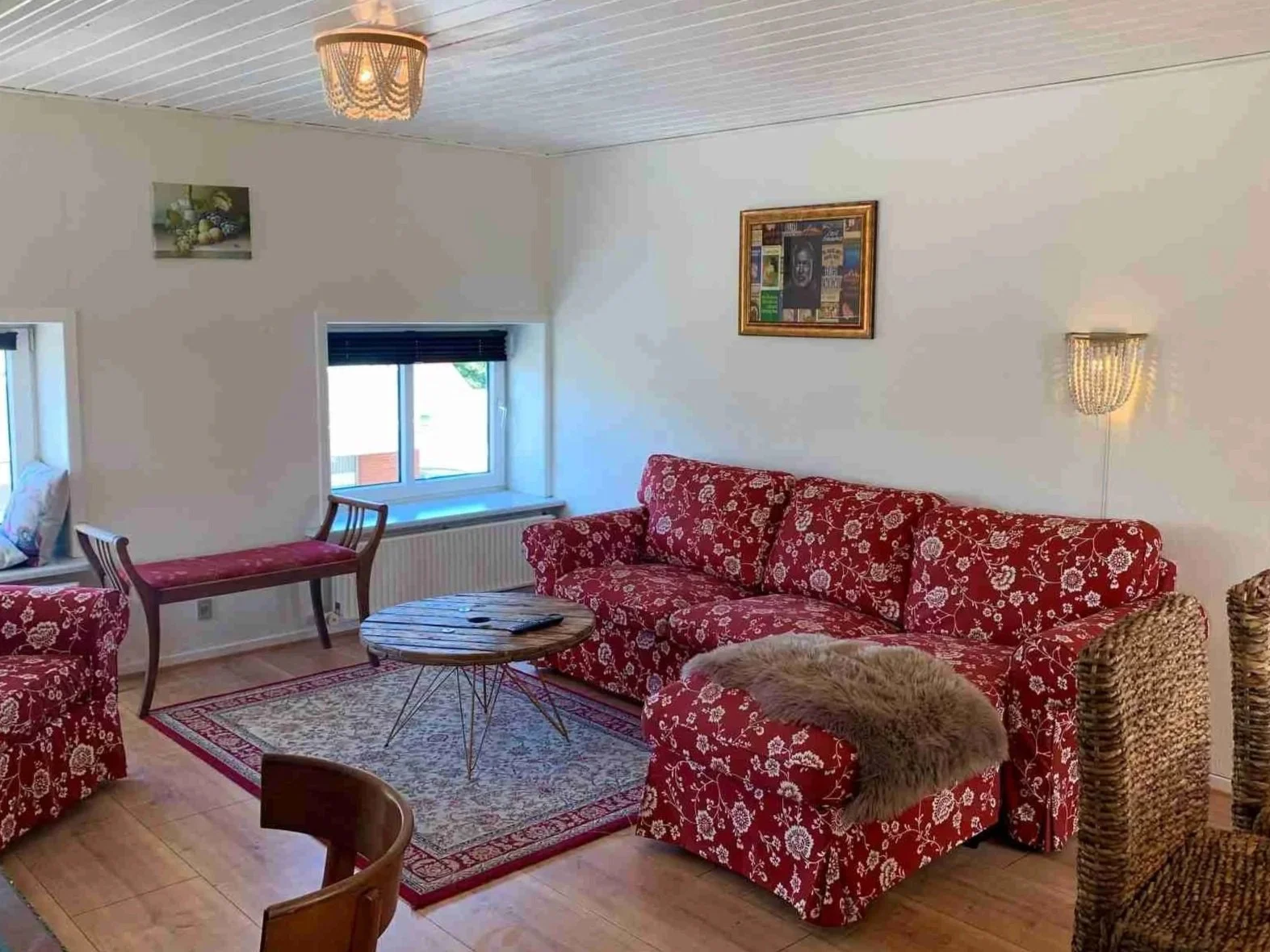 Stue med rød floral sofa, filt på fodende, træbord, vinduer med sorte rullegardiner, vægdekorationer, stor stue med flere siddepladser og blide belysning.