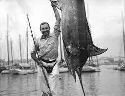 Mand holder en stor fisk foran en marina med bådsejlere i baggrunden.