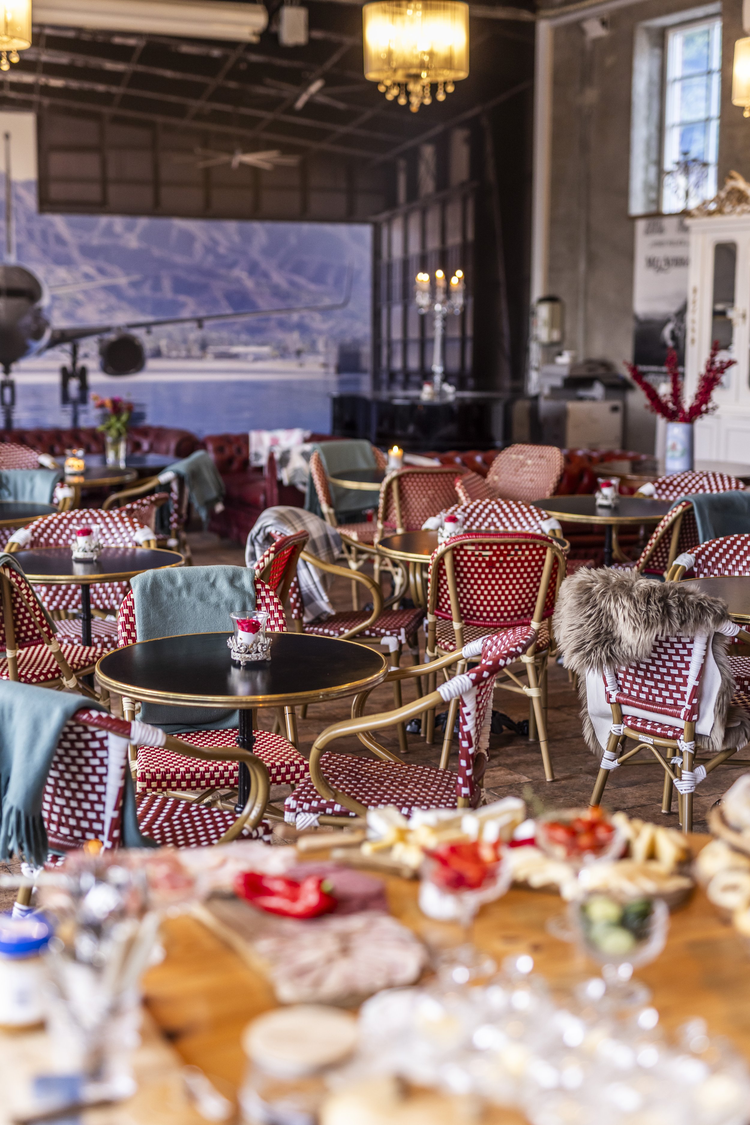 Indendørs restaurant med rød og hvidt mønster på stoleryg og borddækning, lysende lamper, et fly, malerier på væggene og store vinduer, der viser en udsigt over fjeldlandskab.