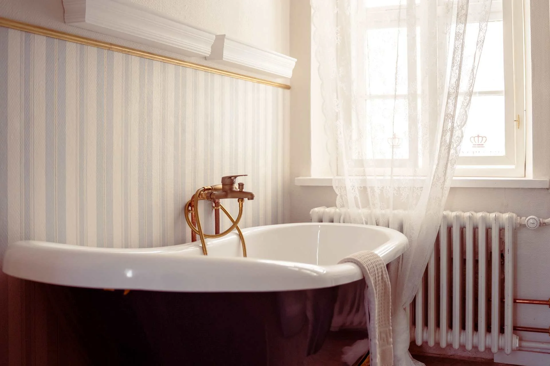 Et badeværelse med et sort badekar, messing armatur, en radiator og et vindue med hvide gardiner.
