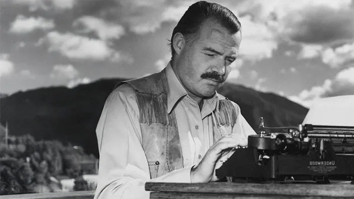 Sort-hvid billede af en mand med sort hår og moustache, der skriver på en skrivemaskine, udenfor med bjerglandskab i baggrunden.