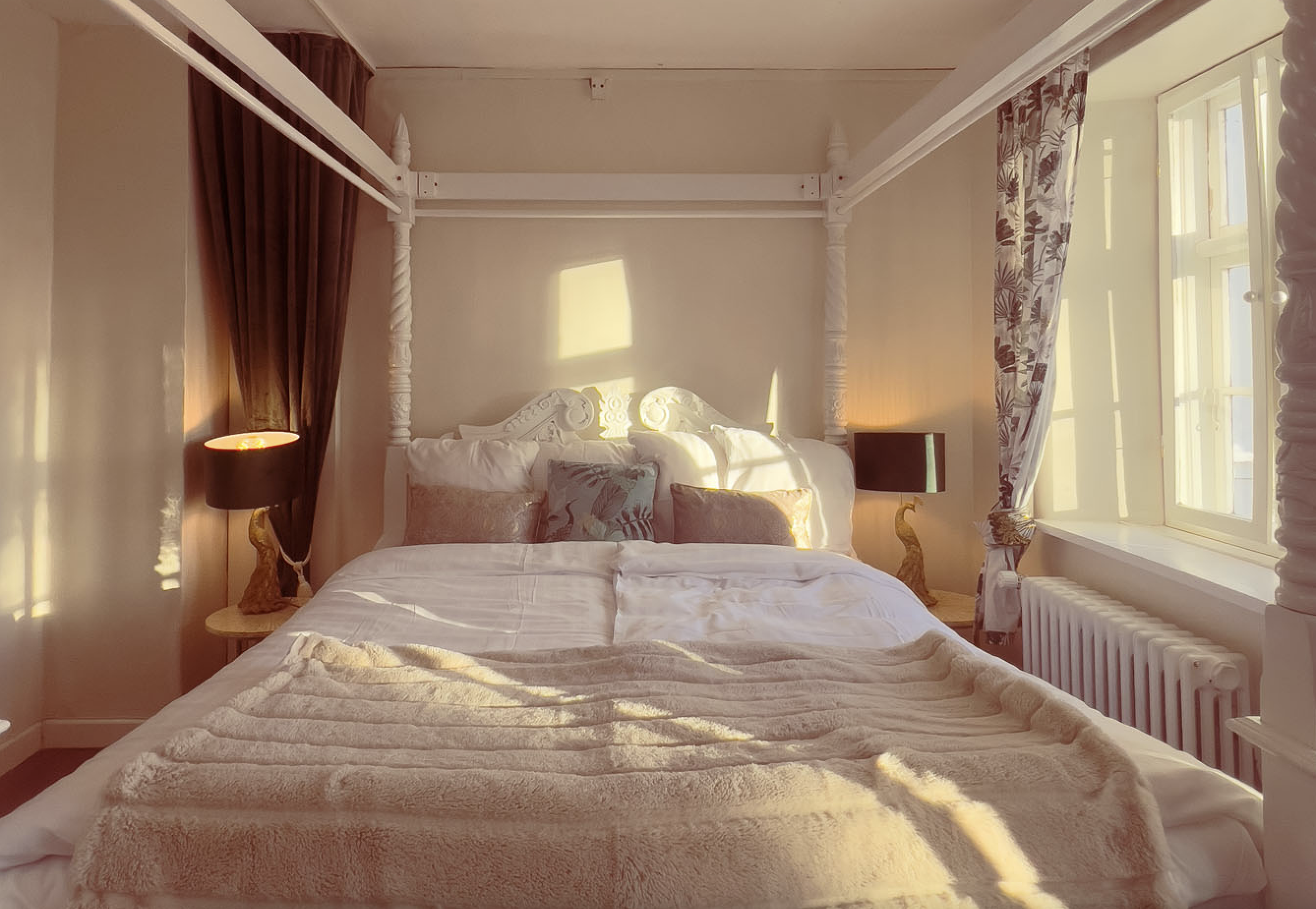 Romantisk soveværelse med himmelseng, puder, og sengetæppe. Stort vindue med aftenlys.