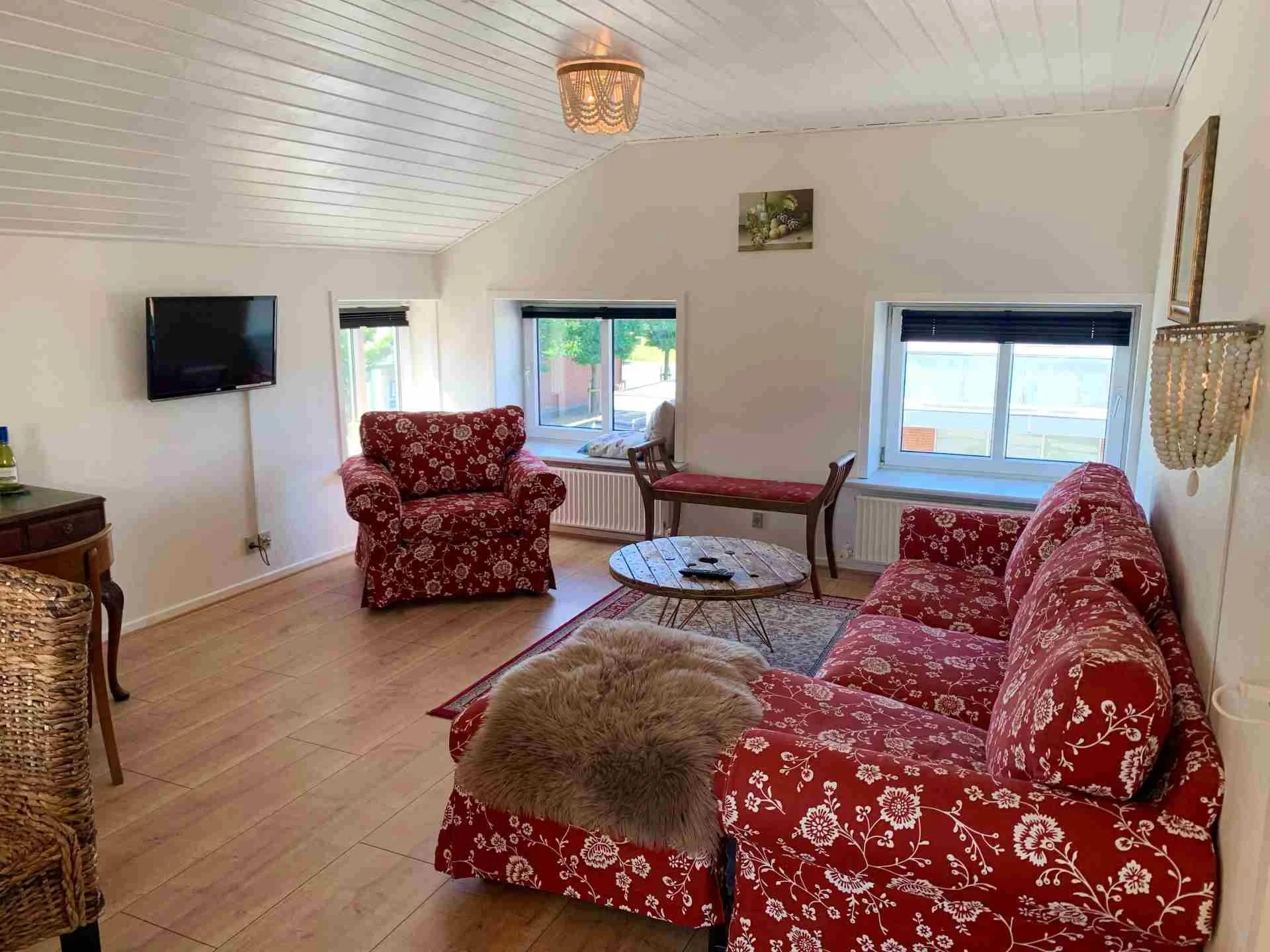 Et hyggeligt stue med tre røde, blomstrede møbler: en sofa, en armchair og et lille bord. Der er tre vinduer med sorte persienner, og en væg med TV og billeder. Der er et tæppe og en plysset pude på sofaen.