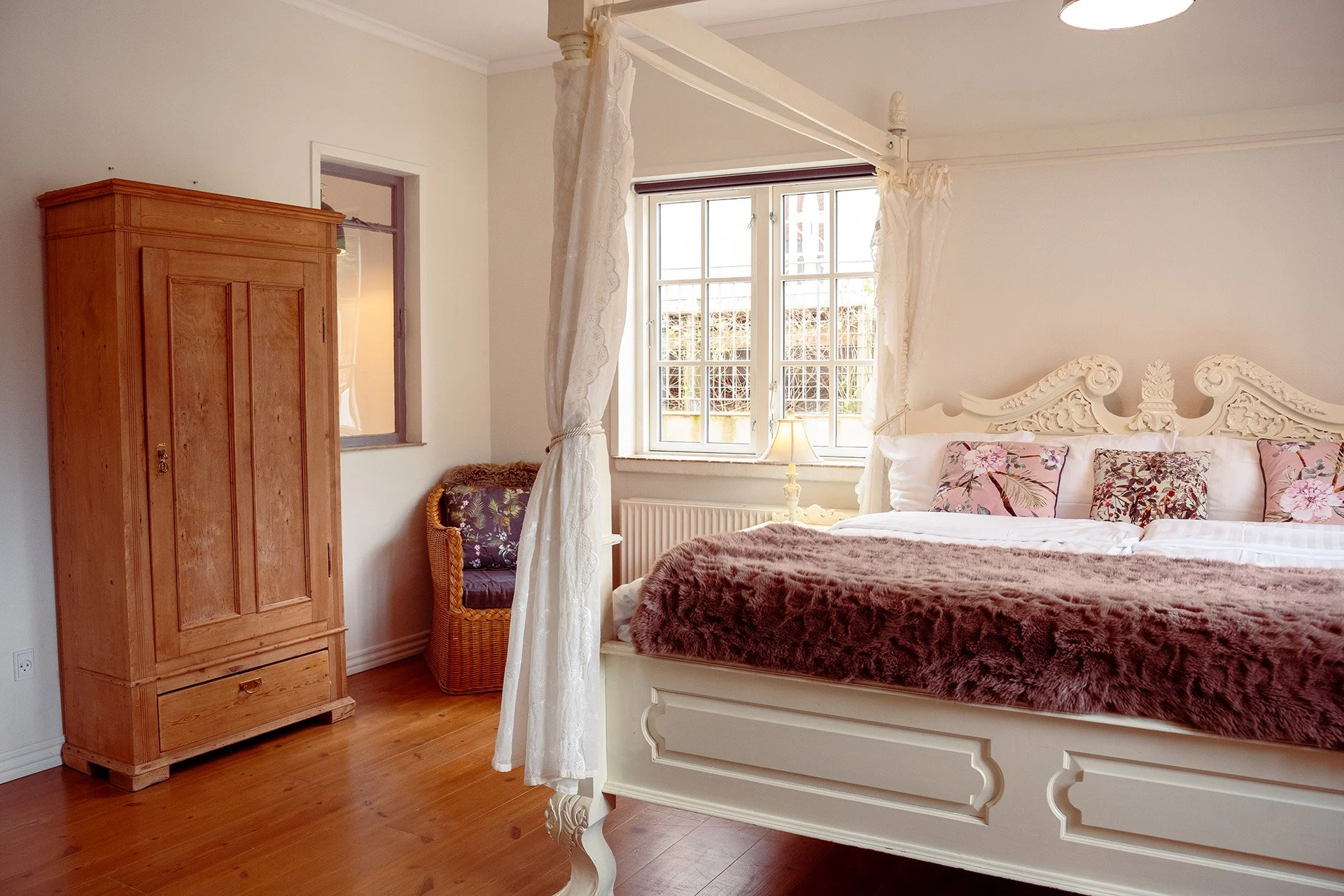 Hvid himmelseng, antikt træskab, rosa puder, pelstæppe