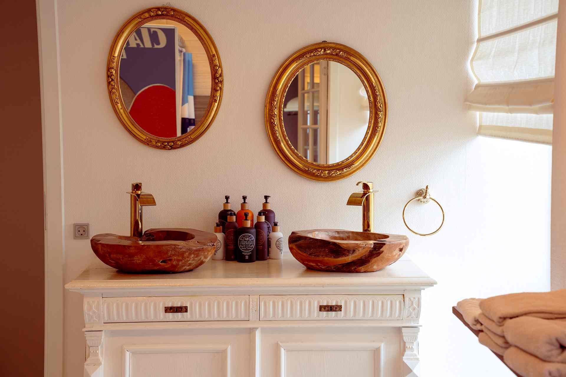 To træbadeværelsesvaser med guldhane og håndklæde på kommode under to gyldne runde vægspiegel.
