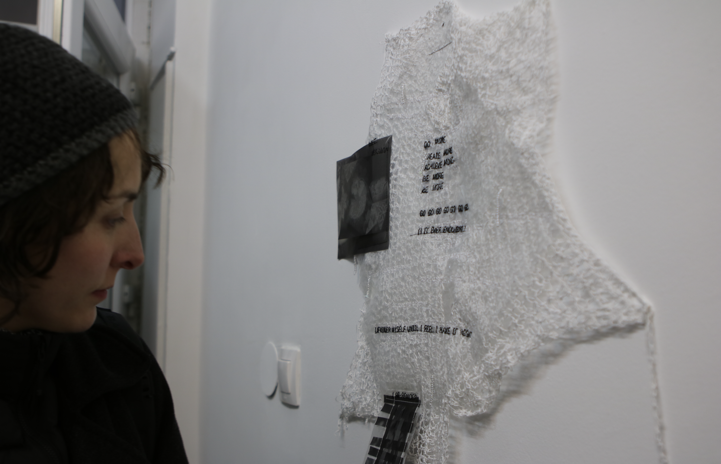 A woman with short hair wearing a knitted hat looks at a textured white art piece with black text and photographs attached to a wall.