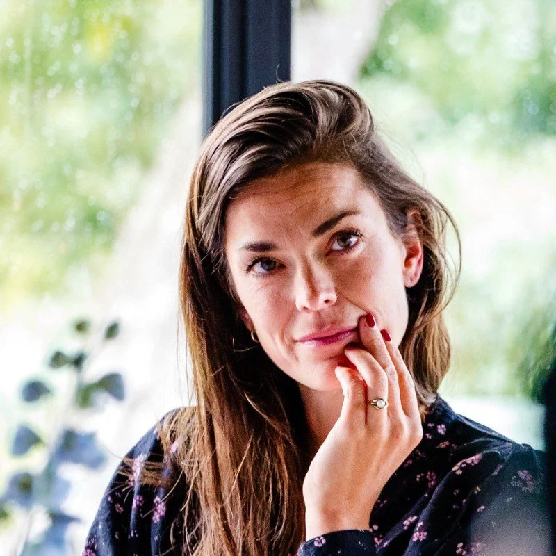 A woman with brown hair and light skin touching her lips near her chin, wearing a dark floral top, standing near a window with blurred greenery outside.
