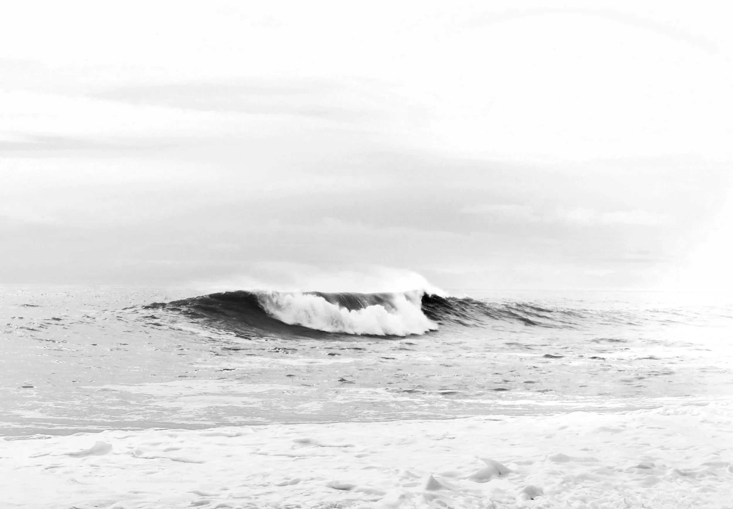 Black and white photograph of a calm ocean with a single wave breaking near the shore under an overcast sky.