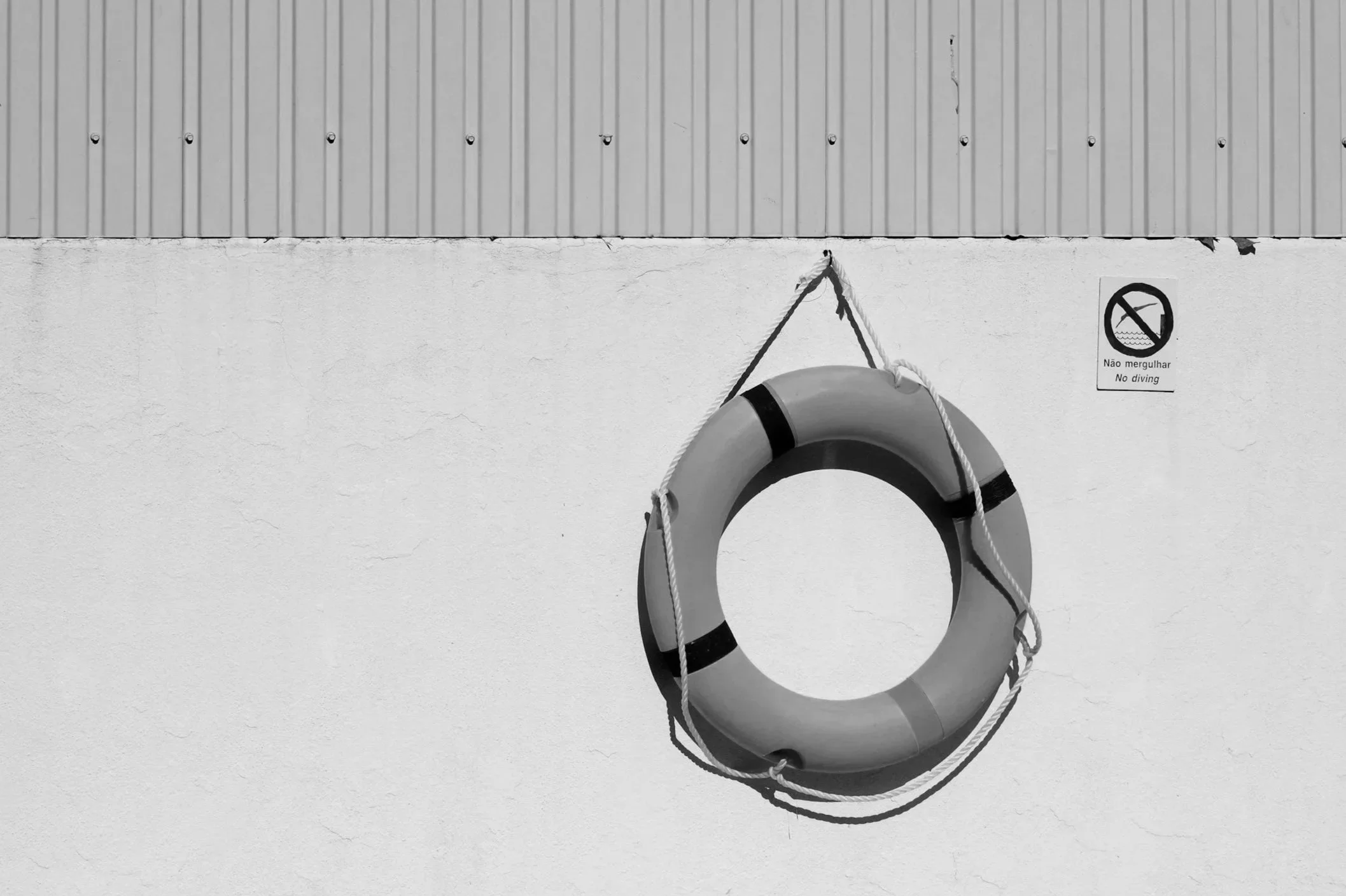 Black and white photo of a life preserver hanging on a white wall, with a small sign to the right that says "No diving" in both English and Portuguese.