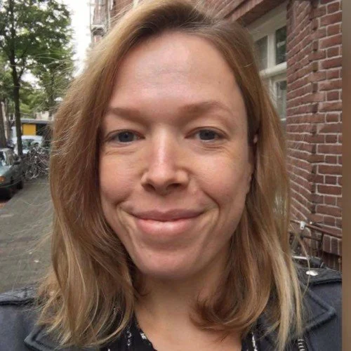 A woman with shoulder-length light brown hair smiling outdoors on a city street with trees and brick buildings in the background.