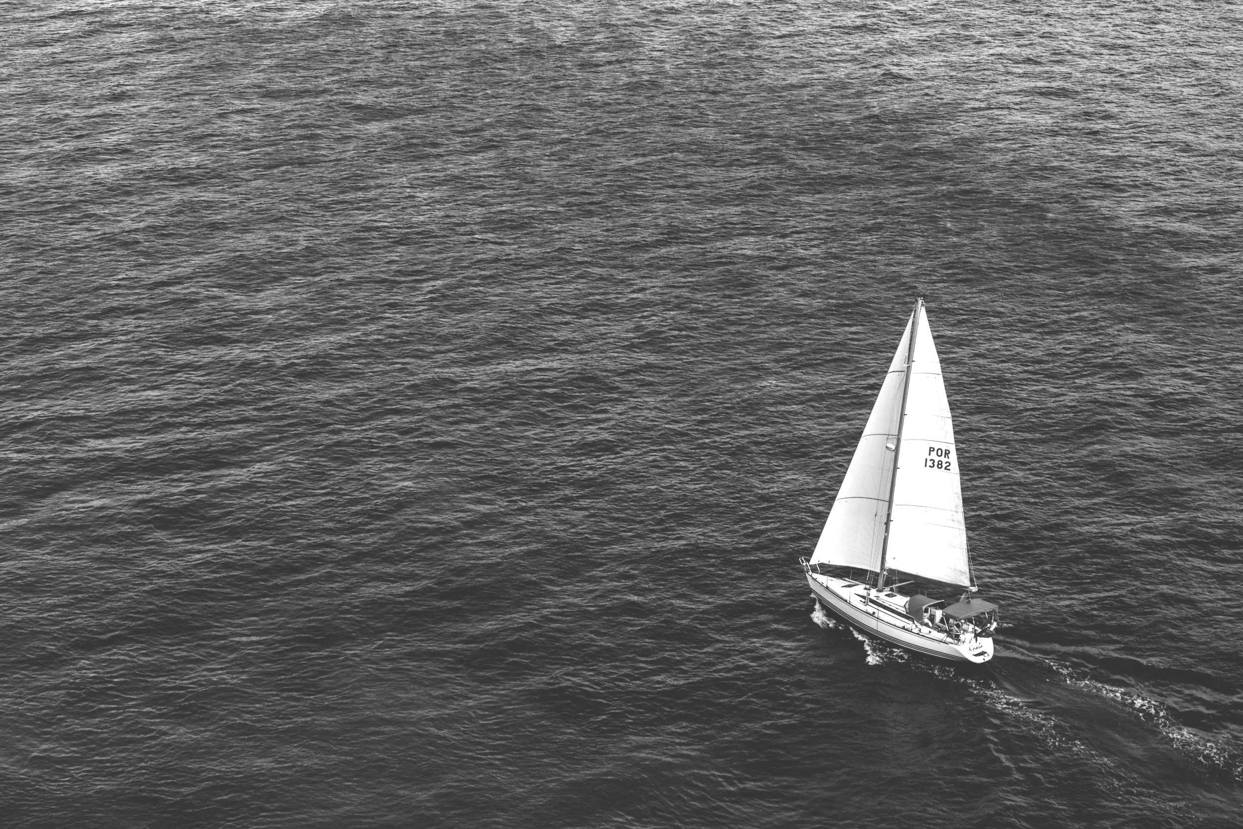 A sailboat on open water with one sail hoisted, moving away from the camera.