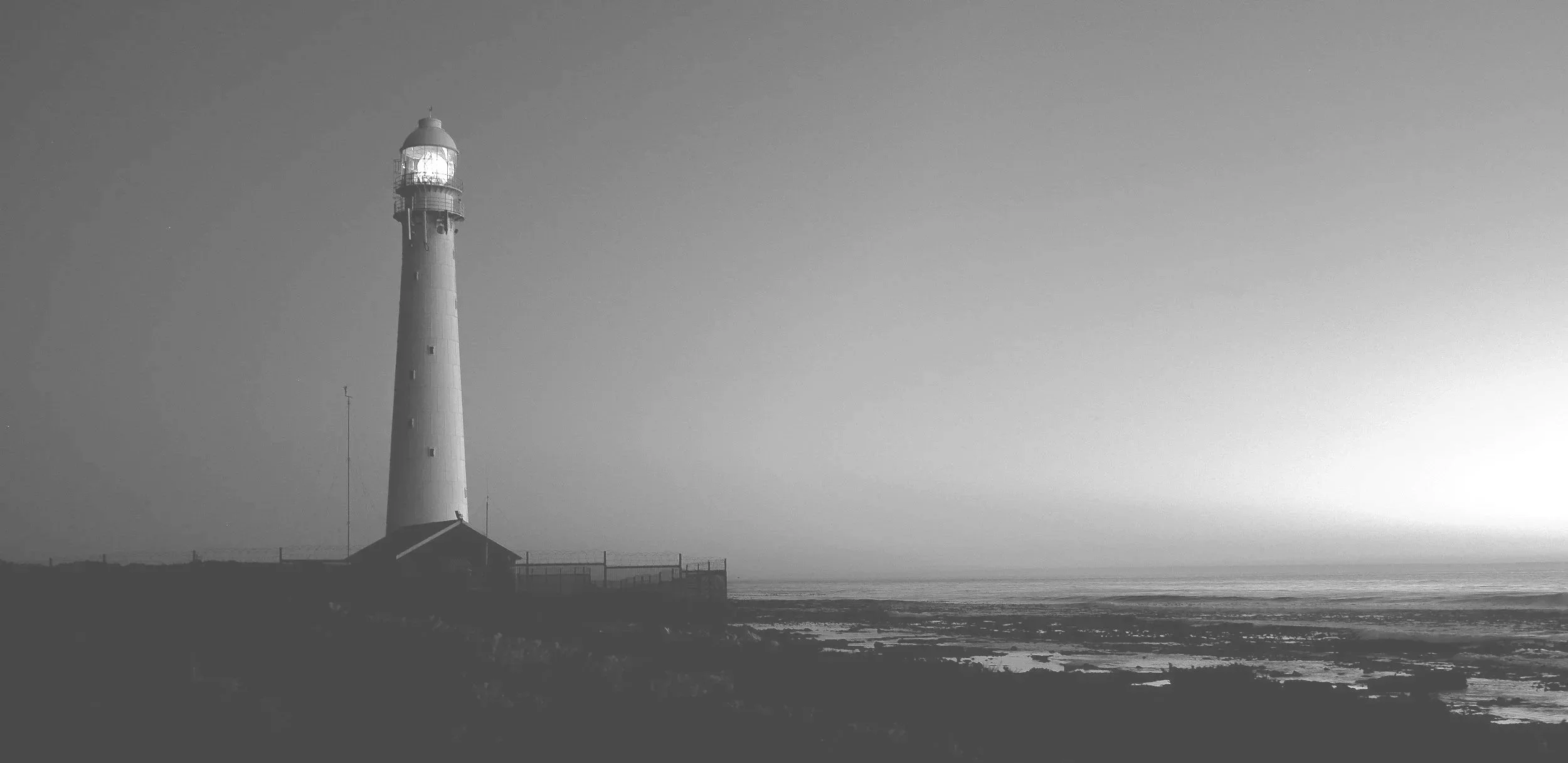A black and white photo of a lighthouse on a coastal area near the ocean, with a small building at its base and a fence around it, under a clear sky.