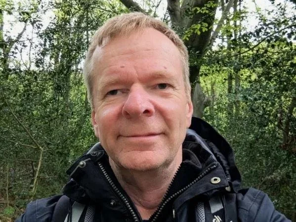 Craig- The owner of Ardent Fireworks, wearing a black jacket with a zipper, standing outdoors among green trees.