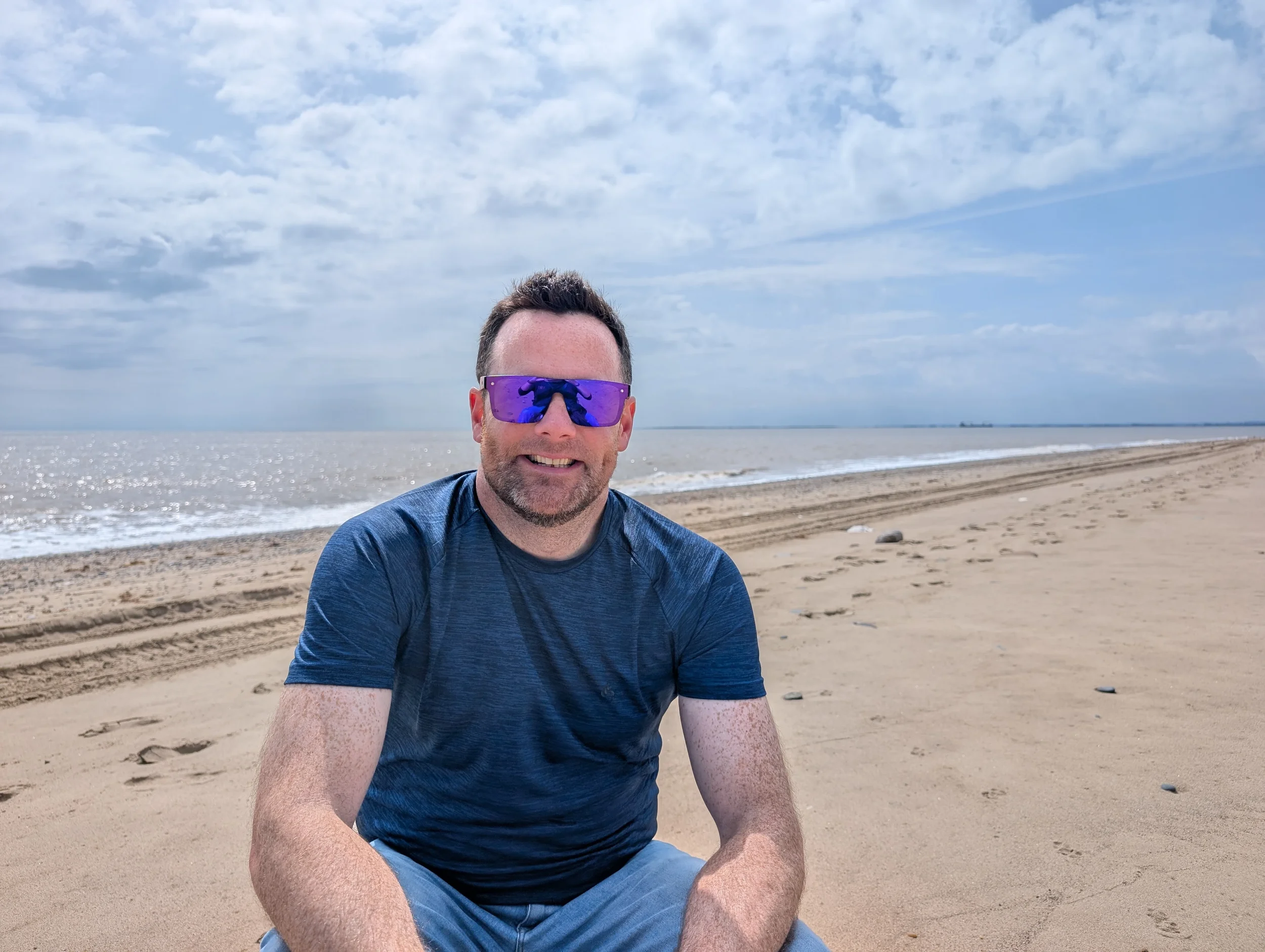 Layne- From Ardent Firworks wearing purple sunglasses and a blue shirt sitting on a sandy beach