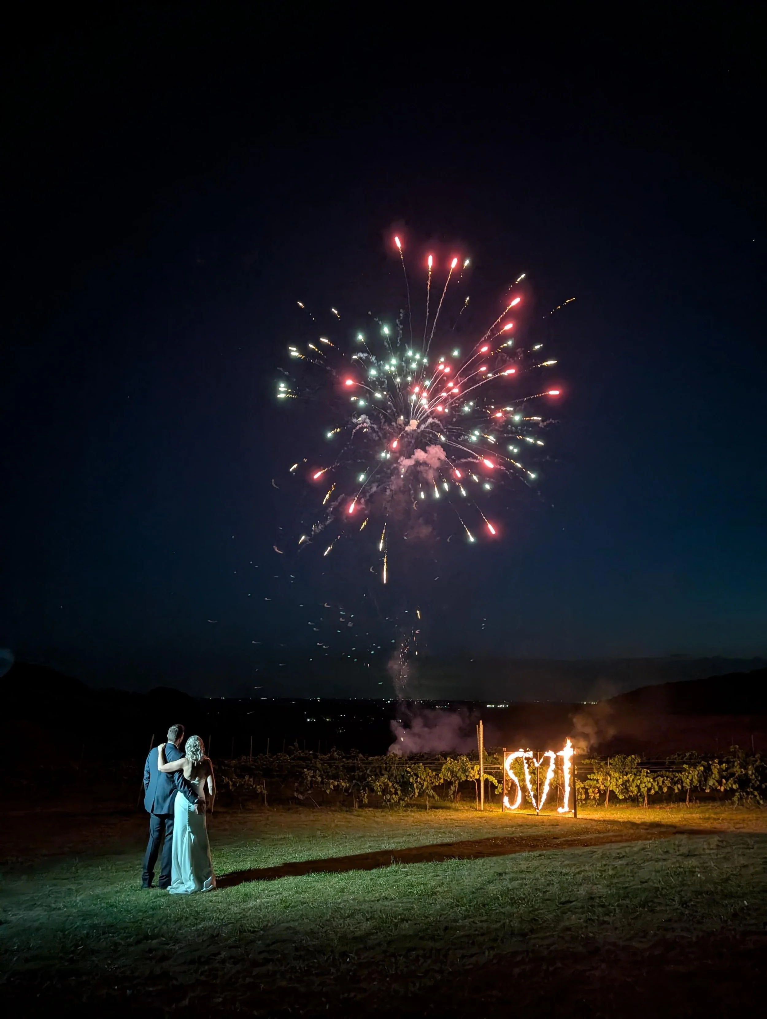 A Wedding couple, standing on grass and watching fireworks in the night sky, with a Fire Rope sign displaying the initials.