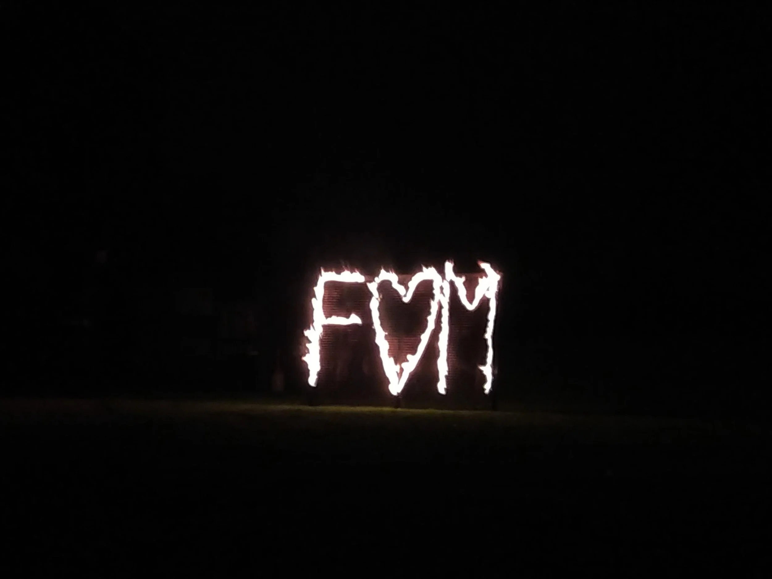 A lit fire art installation spelling out 'F' and 'M' with a heart shape in the middle, against a dark background.