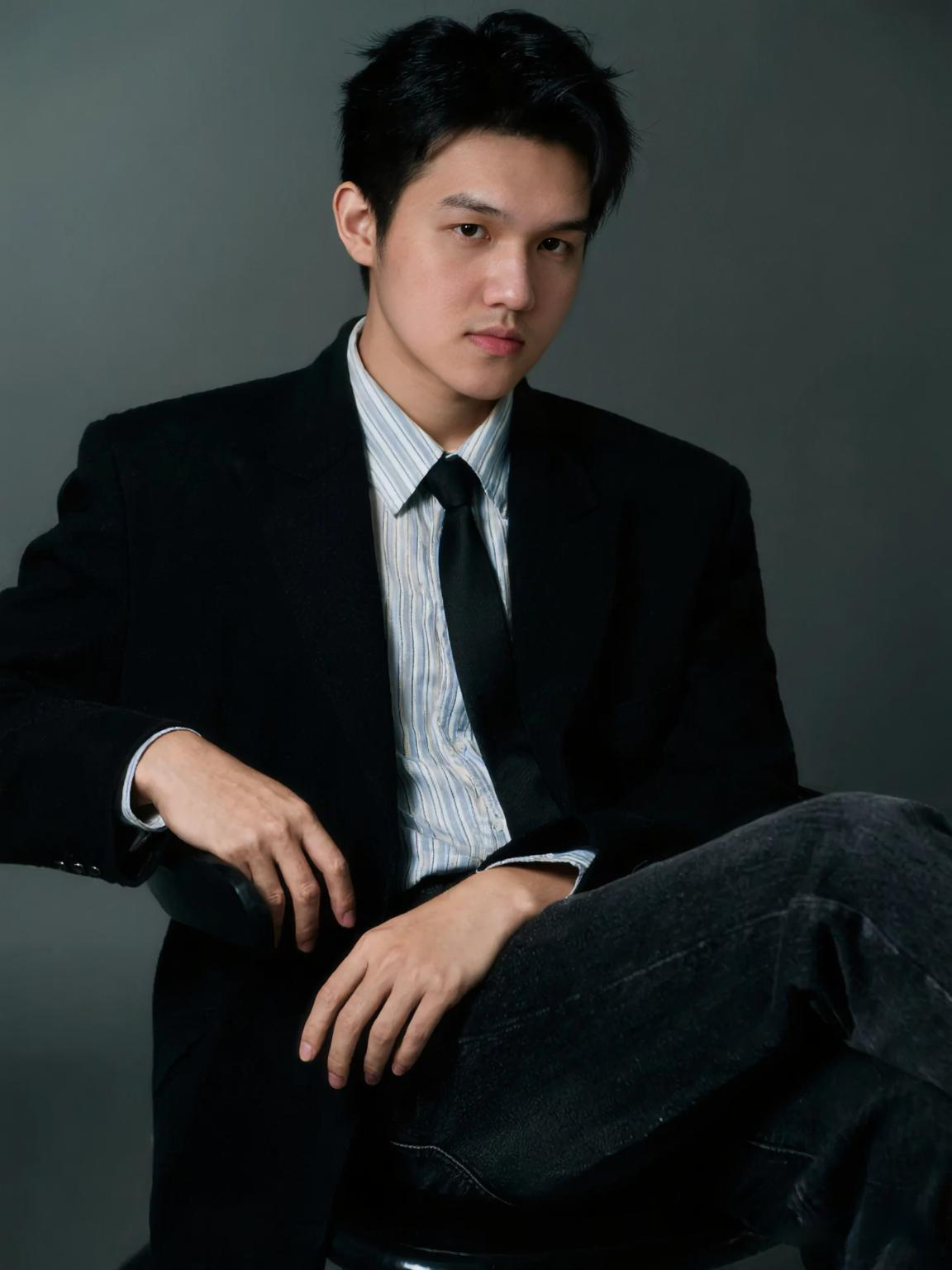 A young man with black hair wearing a black suit, white striped shirt, and black tie, sitting in a dark room with a serious expression.