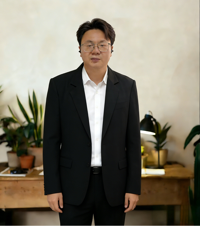 A young man wearing a black suit, white shirt, and glasses standing indoors with a neutral expression. Background includes a wooden table with potted plants and a desk lamp.