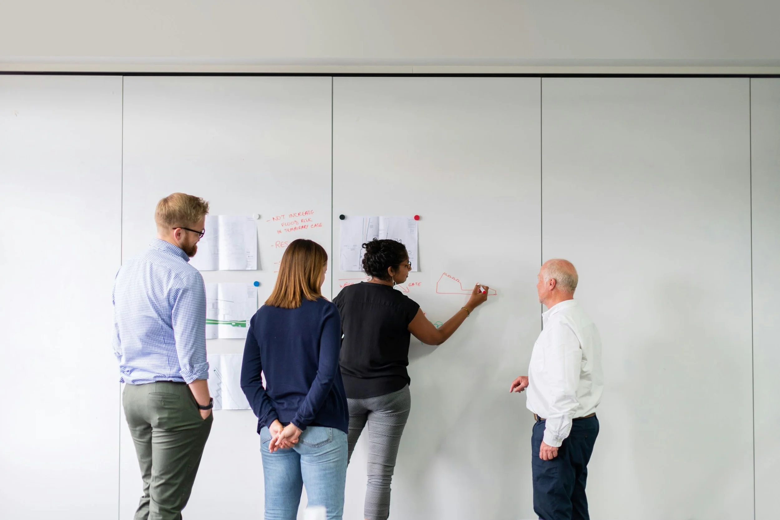 Four people standing in front of a whiteboard, one woman drawing on it with a marker, others observing.