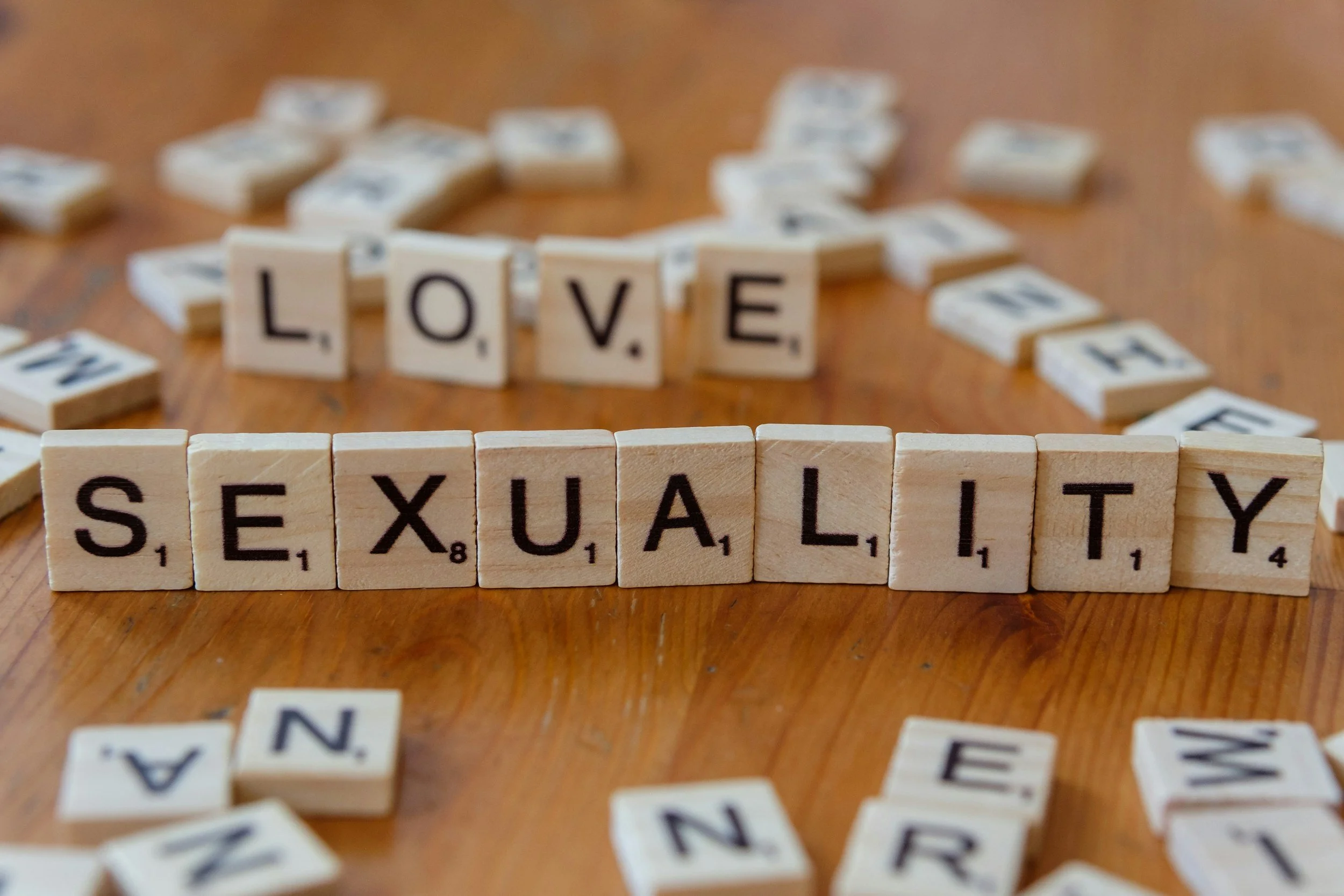 Scrabble-Steine bilden die Worte 'LOVE' und 'SEXUALITY' auf einem Holztisch, umgeben von verstreuten Buchstabensteinen.