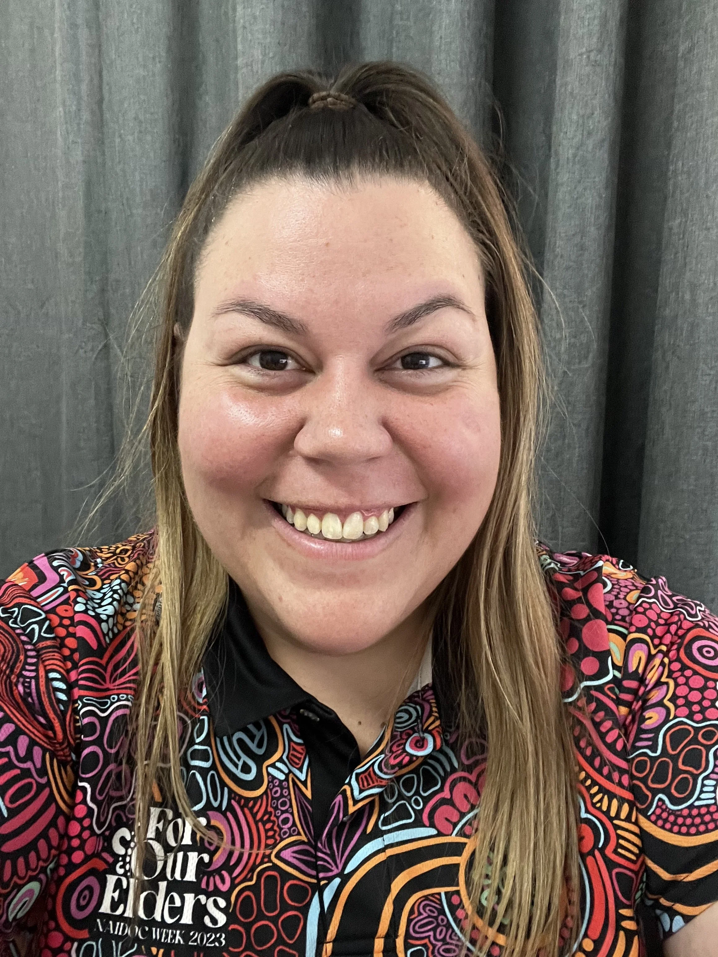 A woman smiling with a colorful shirt that has a logo on it, against a dark curtain background.