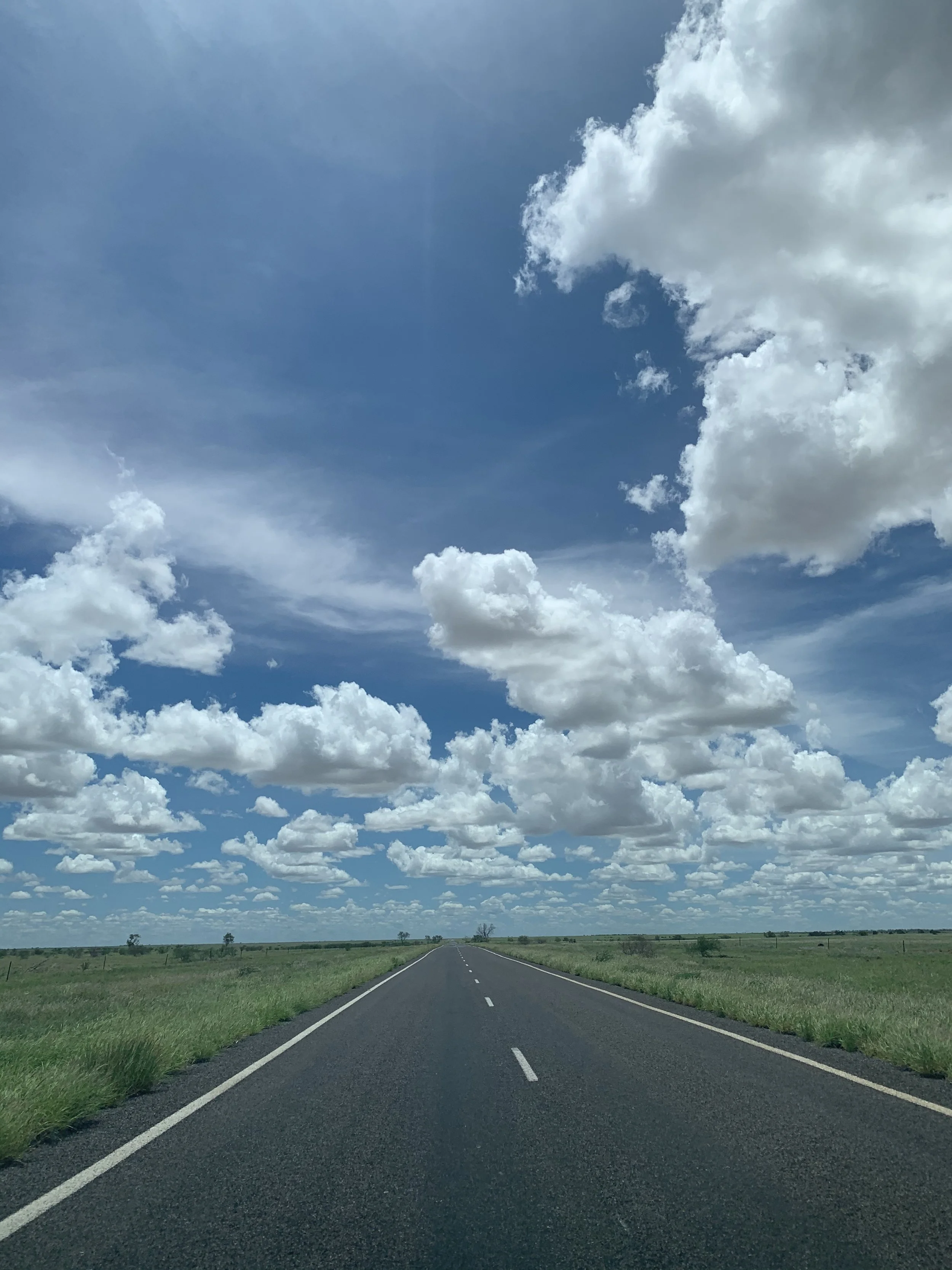 A straight open road stretches into the horizon amidst green fields, under a blue sky filled with white, fluffy clouds.