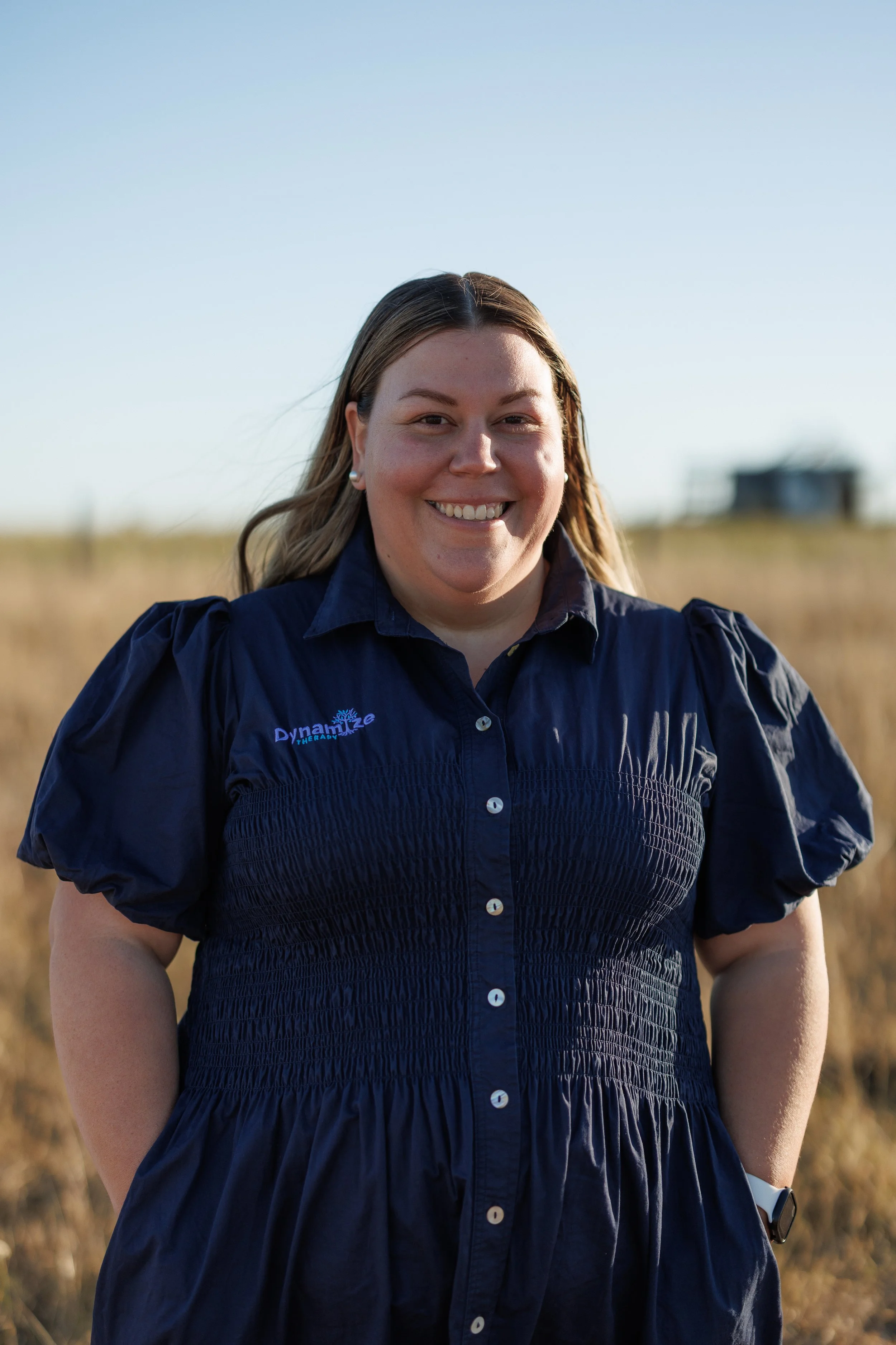 Cass, standing with a smile, outdoors in a field, wearing a navy blue dress with text that says "Dynamike Therapy." The background shows a clear sky and a blurred building in the distance.