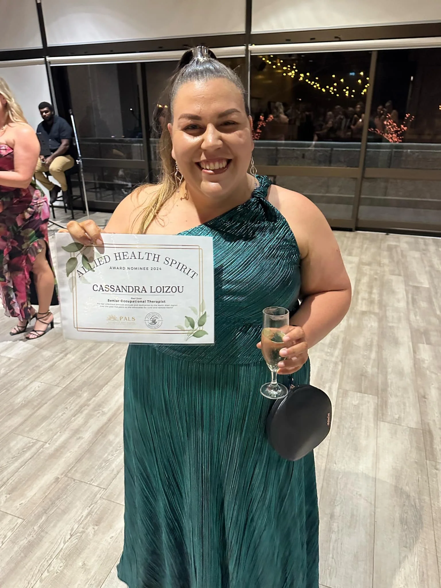 Woman in a green dress holding a certificate and a glass of champagne at an indoor event.