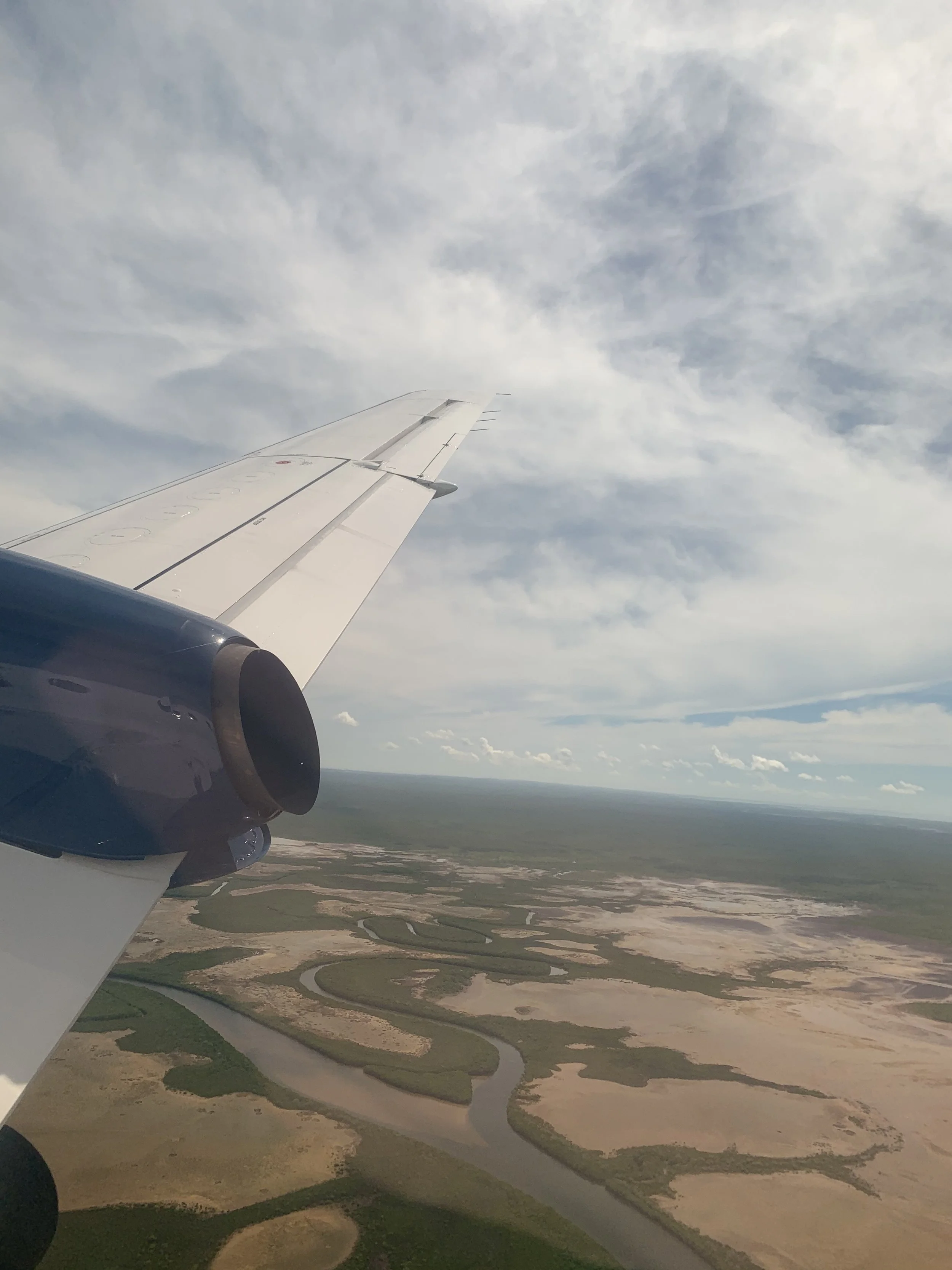 View from an airplane window showing part of the wing, a blue sky with some clouds, and a landscape of winding rivers and wetlands below.