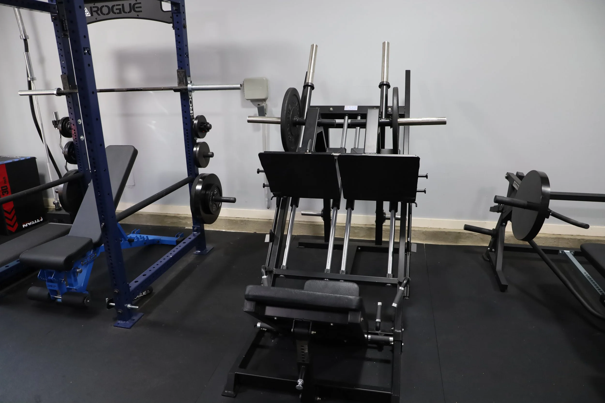 Gym equipment including a blue power rack with barbells and weight plates, a black sled with adjustable weight, and a back extension bench, in a fitness room with a white wall.