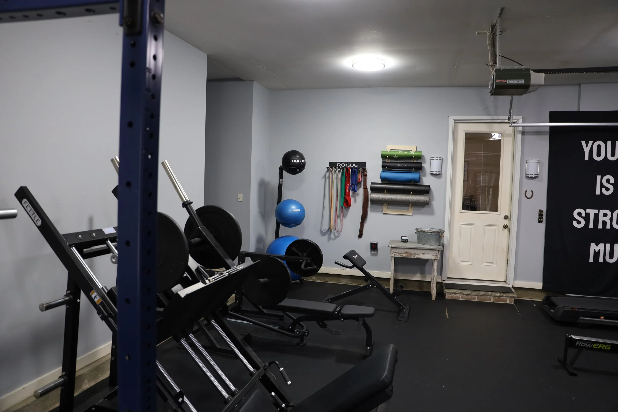 Home gym with weightlifting equipment, exercise balls, resistance bands, and foam rollers against a light blue wall.