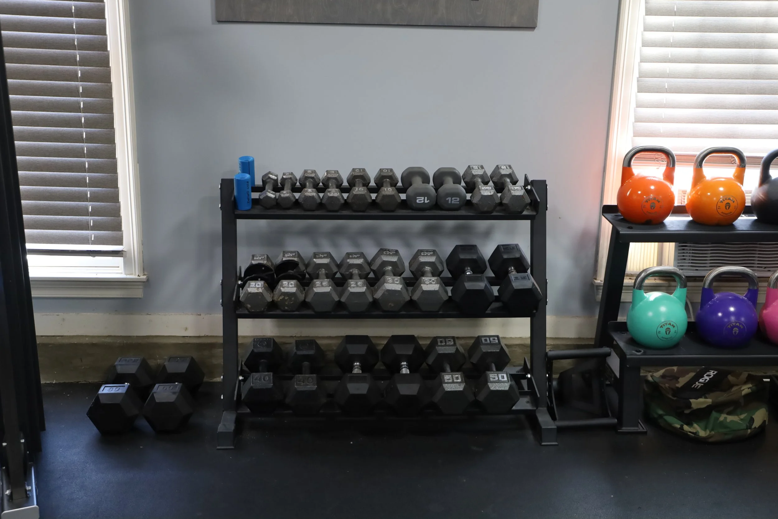 Dumbbells on a black rack with three shelves, a set with blue weights on the top, others with black and gray weights on the middle and bottom shelves, additional dumbbells on the floor. To the right, colorful kettlebells on a black stand near a windo