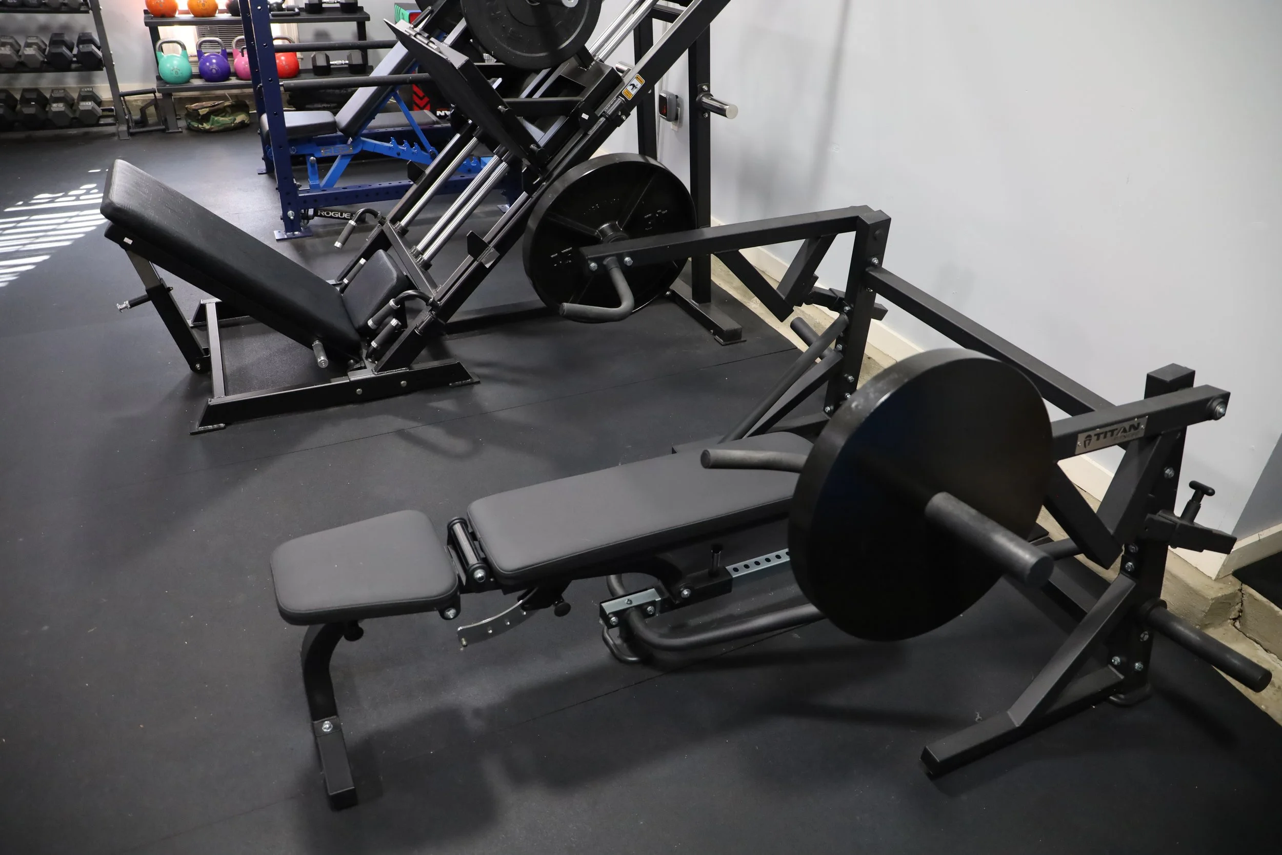 Gym equipment including a leg press machine and an adjustable weight bench with weight plates on a black rubber floor.