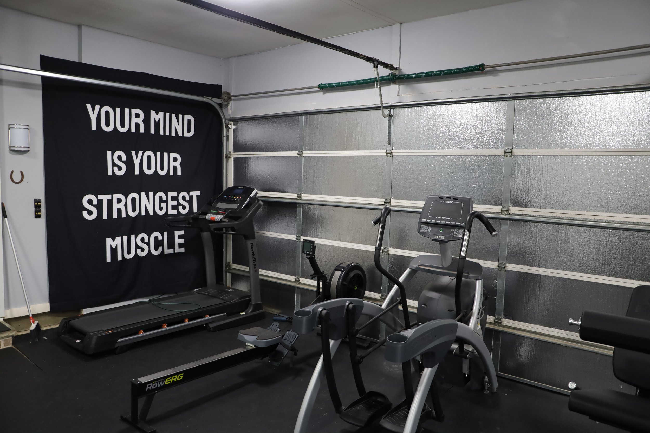 Home gym with treadmill, elliptical machine, rowing machine, and exercise bench inside a garage. A black curtain with white text reads, "Your mind is your strongest muscle."