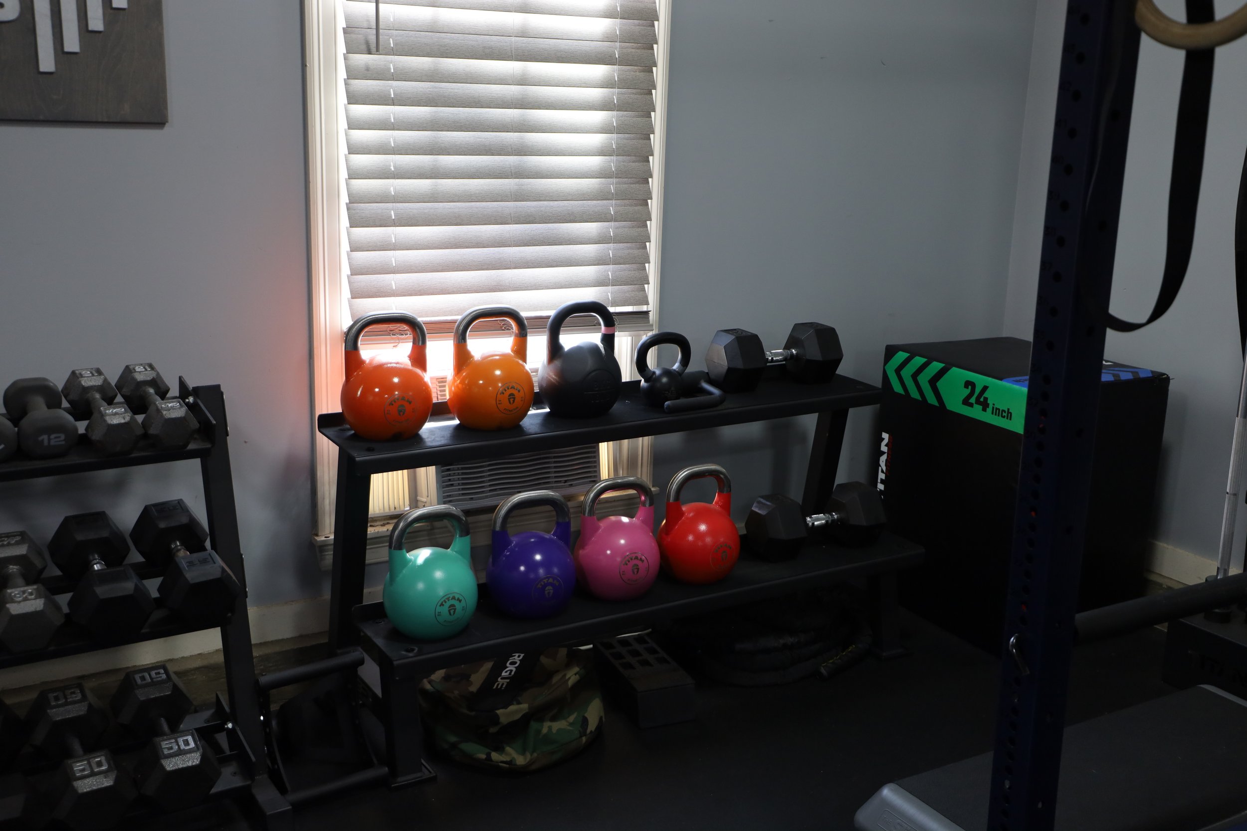 Organized home gym equipment including colorful kettlebells, dumbbells, and a storage rack near a window with closed blinds.