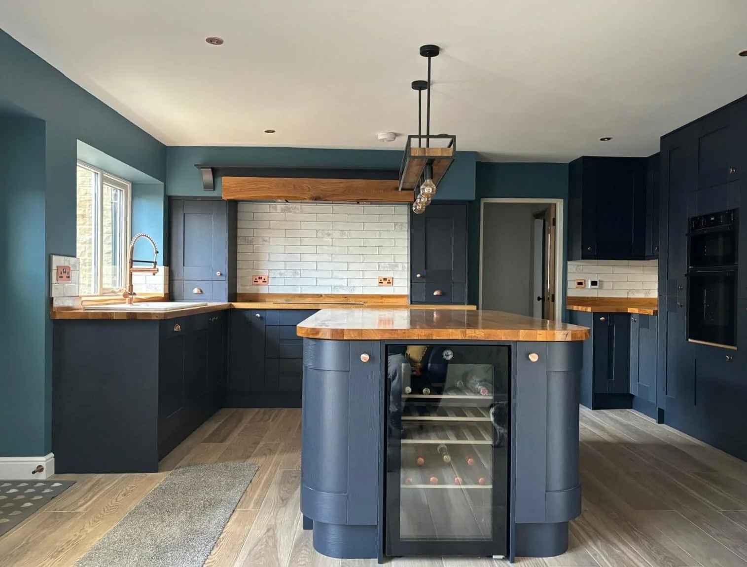 Modern kitchen with navy blue cabinets, wooden countertops, a small wine cooler, and a brick backsplash. There is a window above the sink, a pendant light fixture, and a doorway.