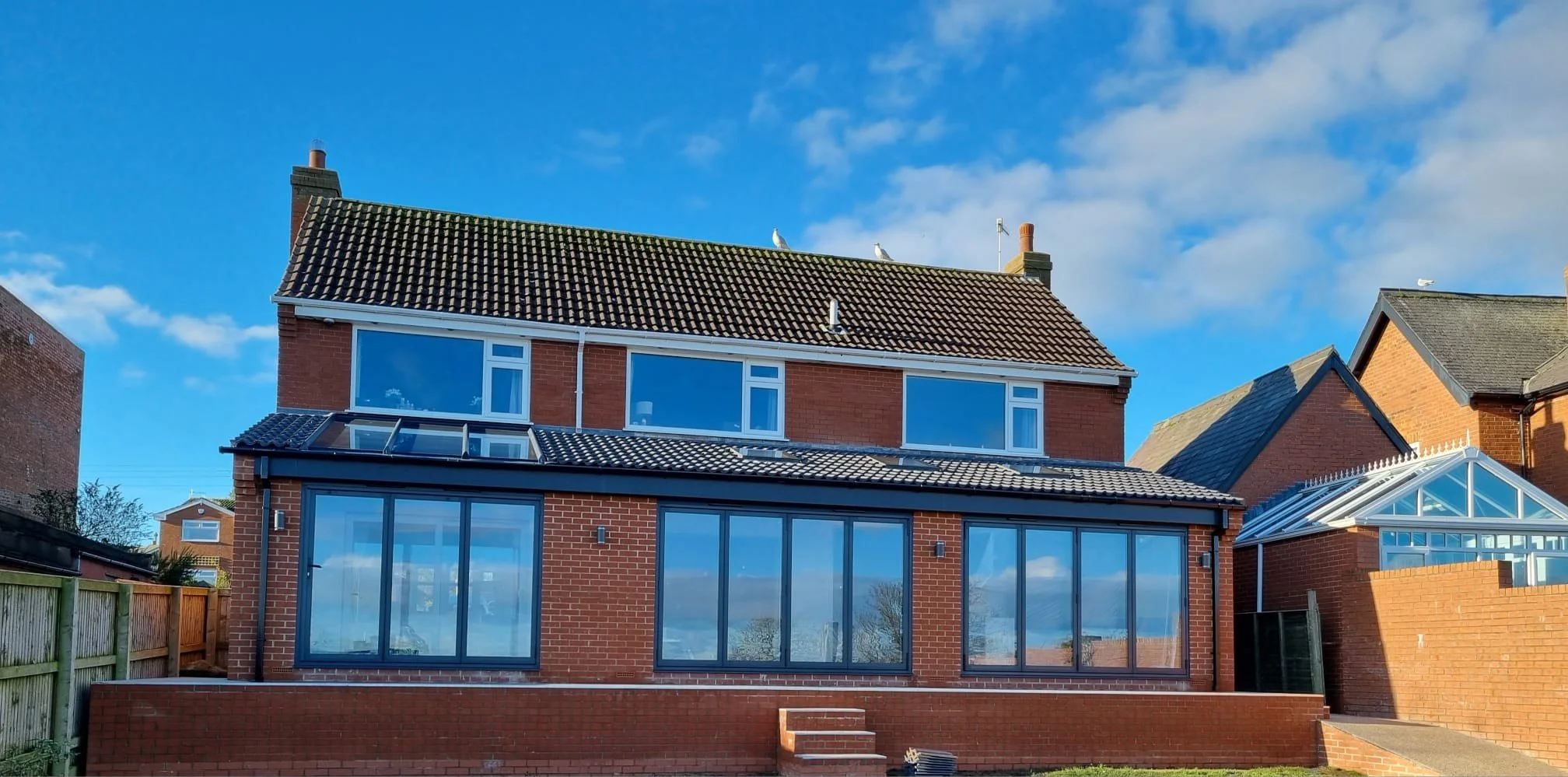 A three-story brick house with a glass extension and a tiled roof, set against a blue sky with a few clouds.