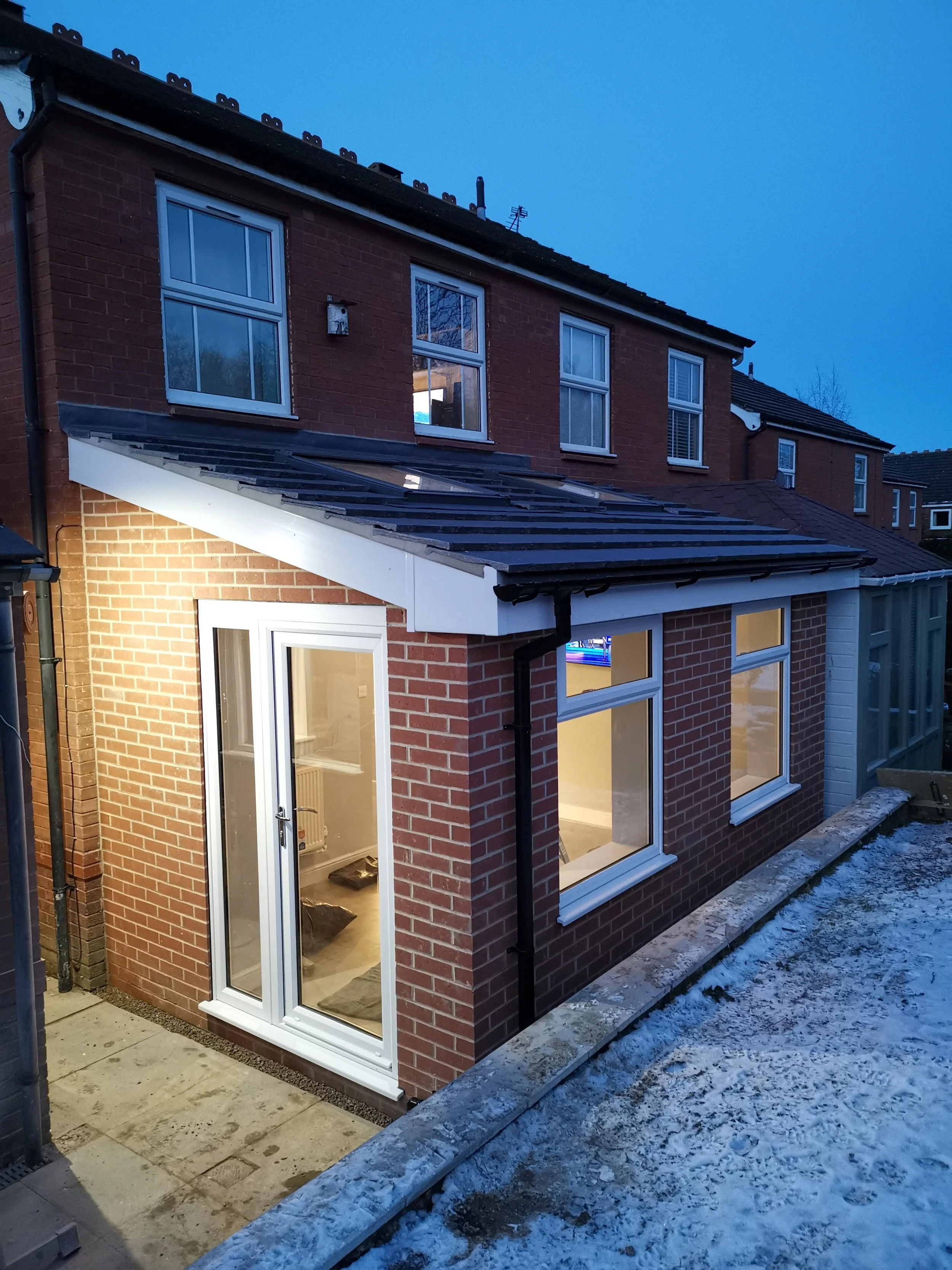 A two-story brick house with a small extension at the back, featuring white-framed glass doors and windows. The area is illuminated with outdoor lighting, and there is snow on the ground.