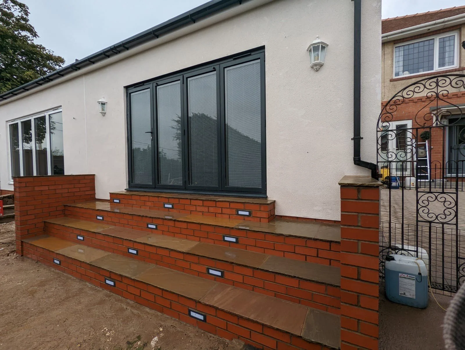 Brick stairs with built-in lighting lead up to a house with large glass doors and beige walls. There is an outdoor wall light and a black downspout visible. Part of another house is visible in the background.